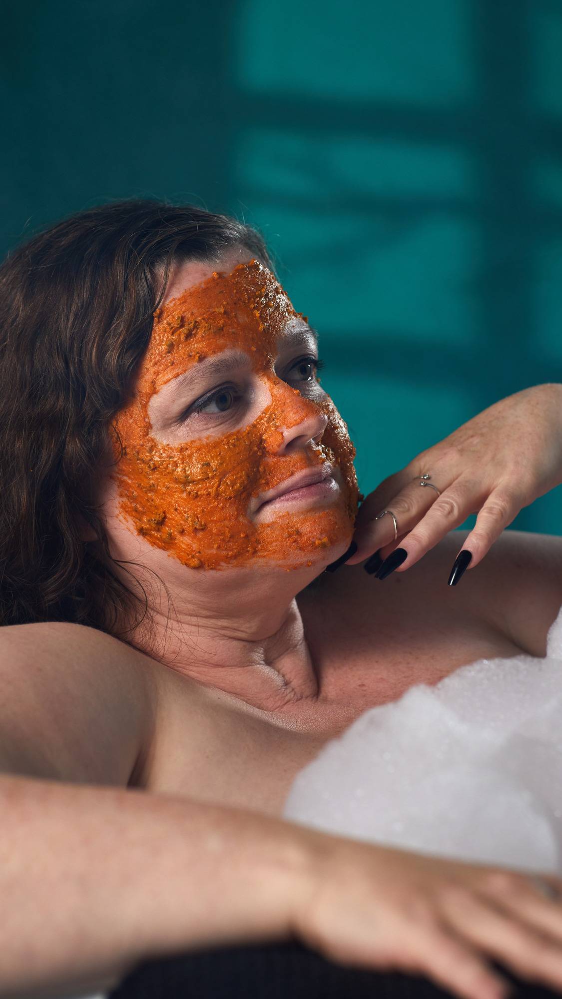 The image shows a person with brown hair relaxing in a bath. A vibrant orange 'Mr. Pumpkin Face' mask has been applied to their whole face. White bath bubbles and a hand touching their face are visible, against a dark teal background.