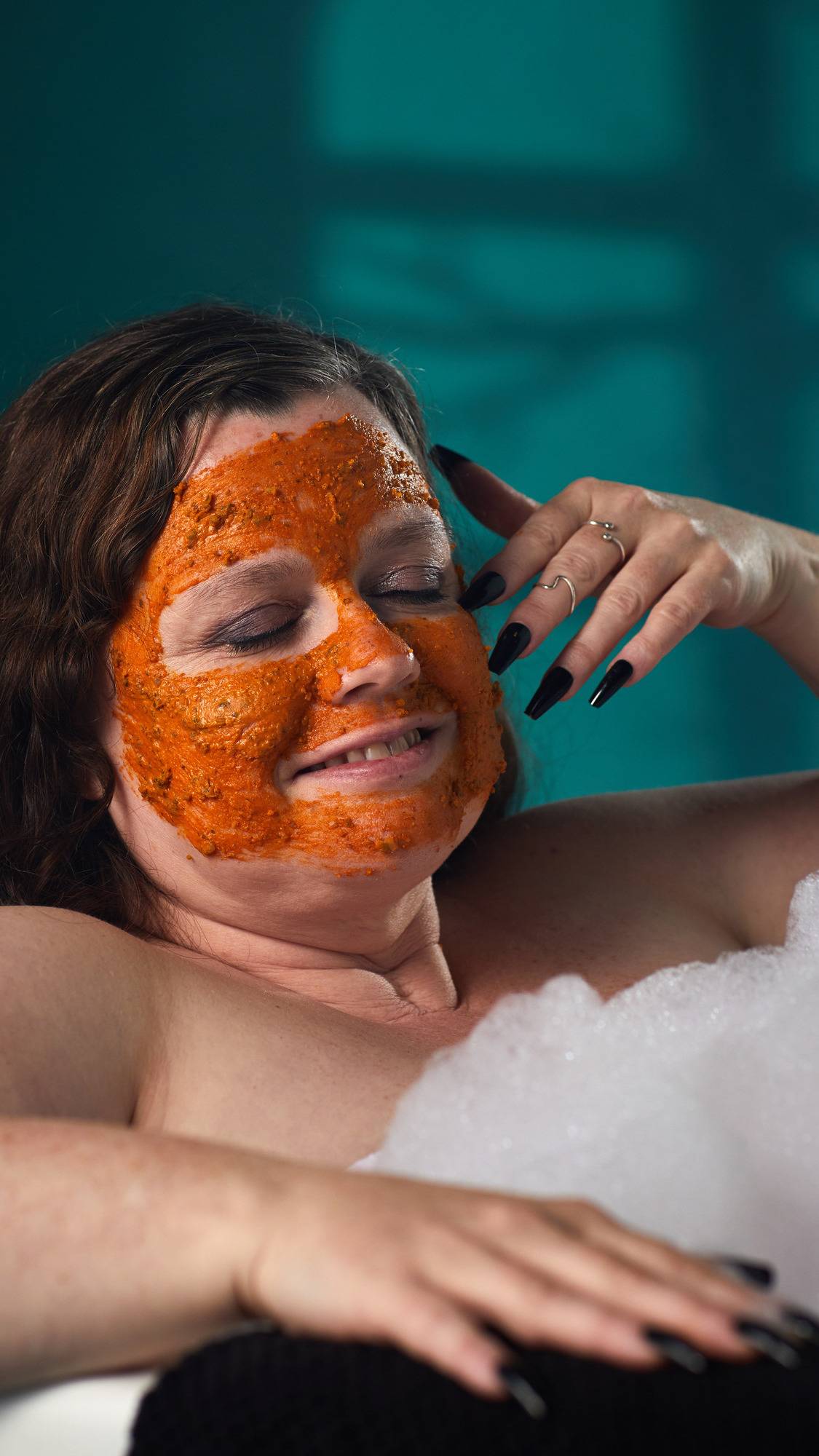 The image shows a person with brown hair relaxing in a bath. A vibrant orange 'Mr. Pumpkin Face' mask has been applied to their whole face. White bath bubbles and a hand touching their face are visible, against a dark teal background.