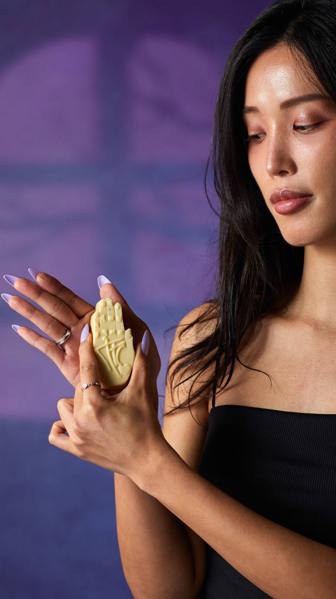 The image shows a person with long dark hair, holding a pale cream, hand-shaped Palm Reader solid hand cream. The product, with its embossed palmistry lines, is held by hands with light purple nails and rings, against a purple background.
