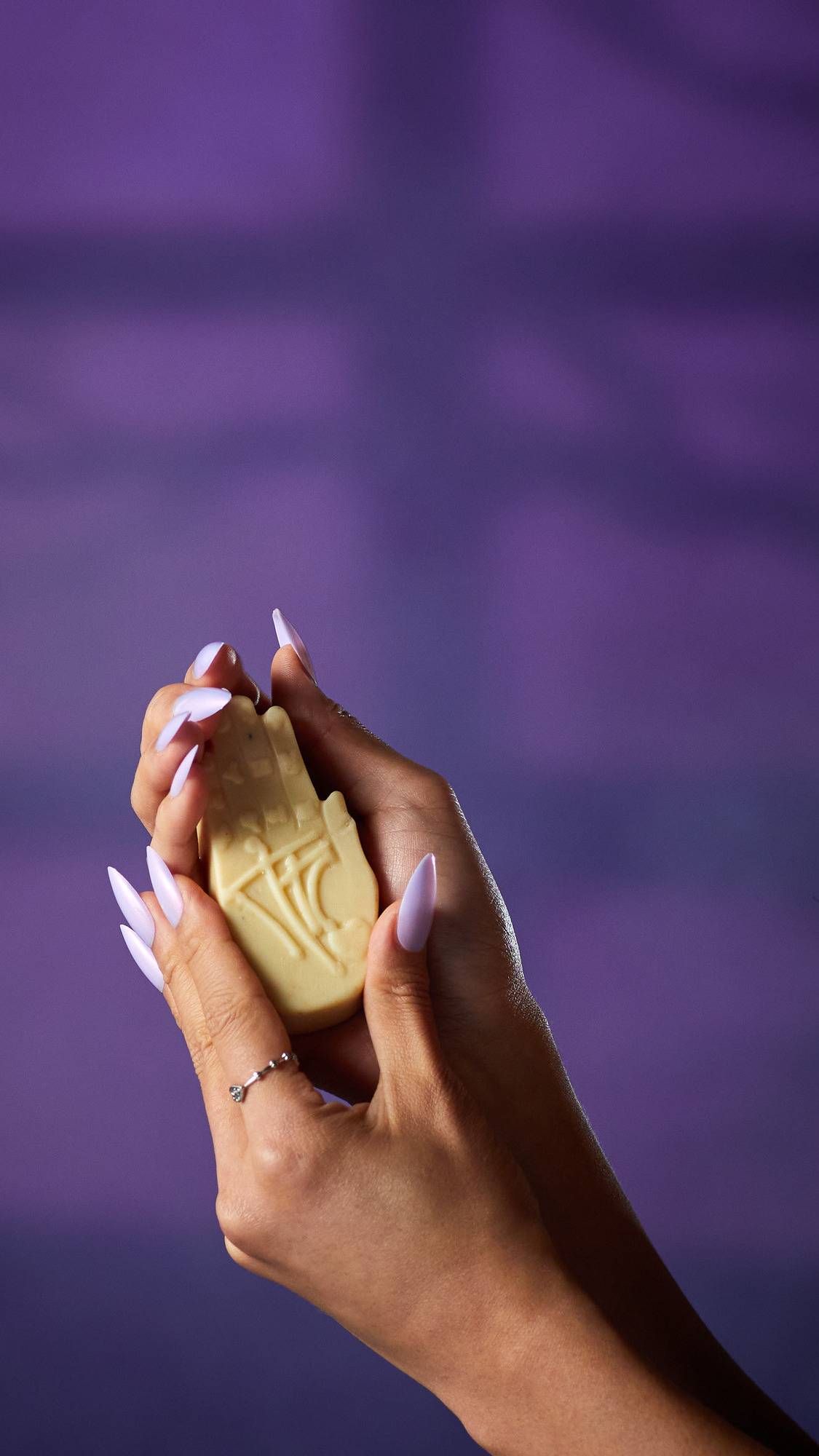 The image shows a pair of hands with long, light purple nails, holding a pale cream, hand-shaped Palm Reader solid hand cream. The product's embossed palmistry lines are visible against a purple background.