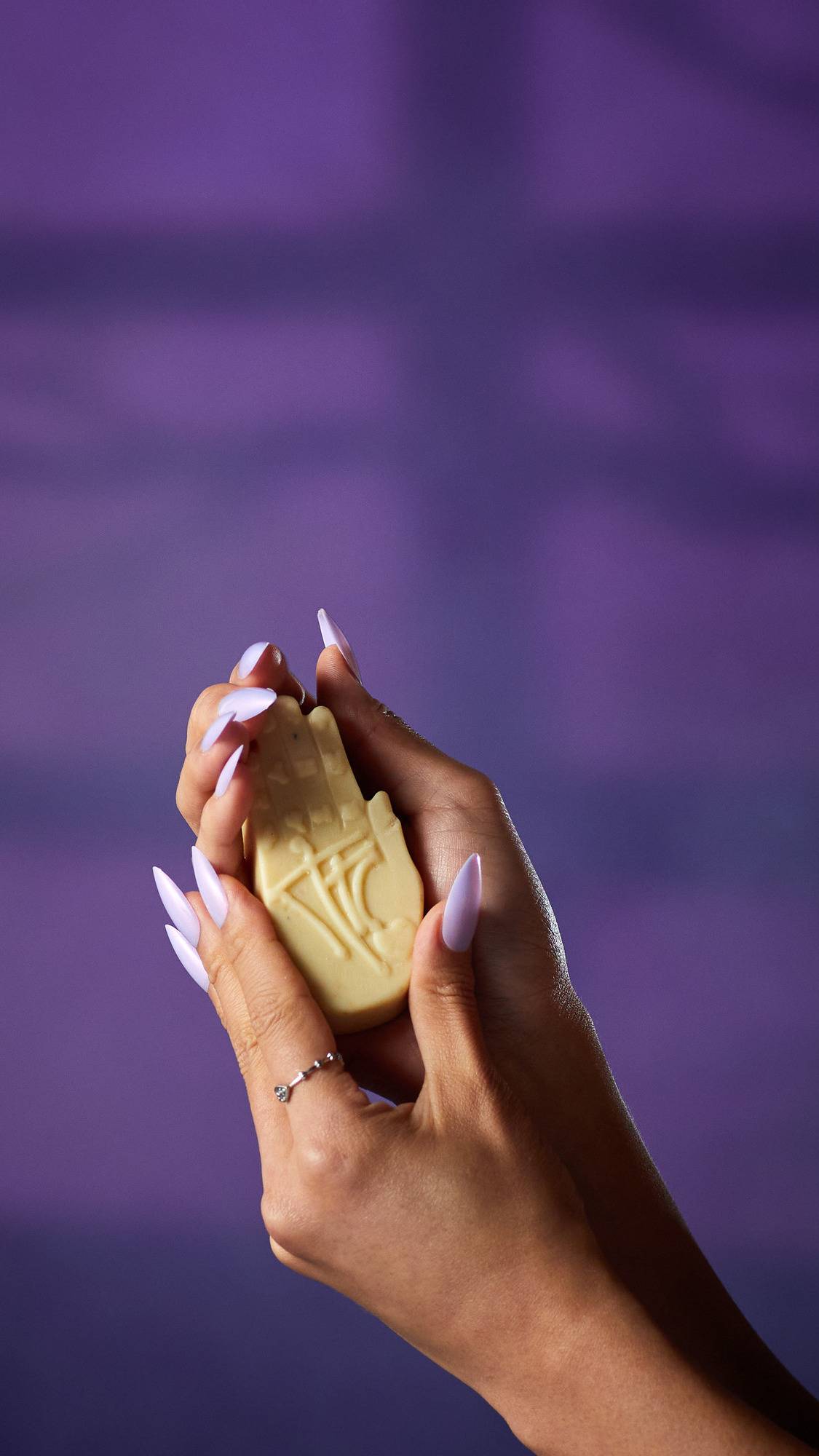 The image shows a pair of hands with long, light purple nails, holding a pale cream, hand-shaped Palm Reader solid hand cream. The product's embossed palmistry lines are visible against a purple background.