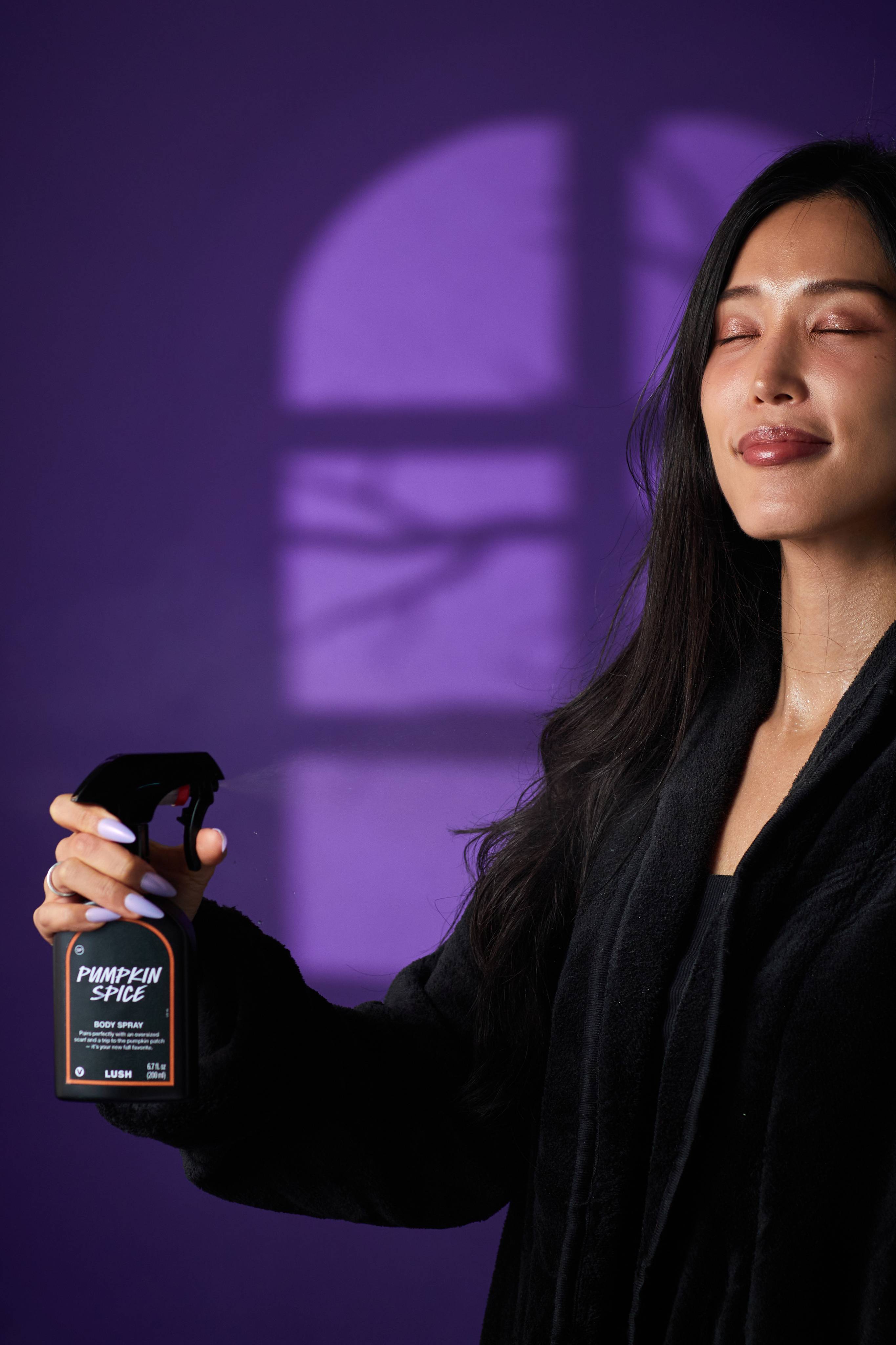 The image shows a person with a serene expression, spritzing a black LUSH 'Pumpkin Spice' body spray bottle on themselves. A hand with light purple nails holds the orange-rimmed label bottle against a purple background with shadow patterns.