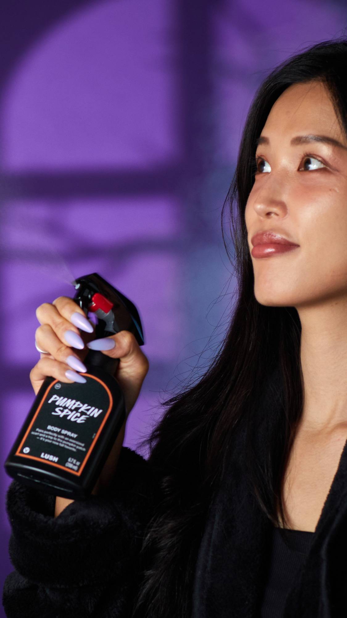 The image shows a person with a serene expression, spritzing a black LUSH 'Pumpkin Spice' body spray bottle. A hand with light purple nails holds the orange-rimmed label bottle against a purple background with shadow patterns.