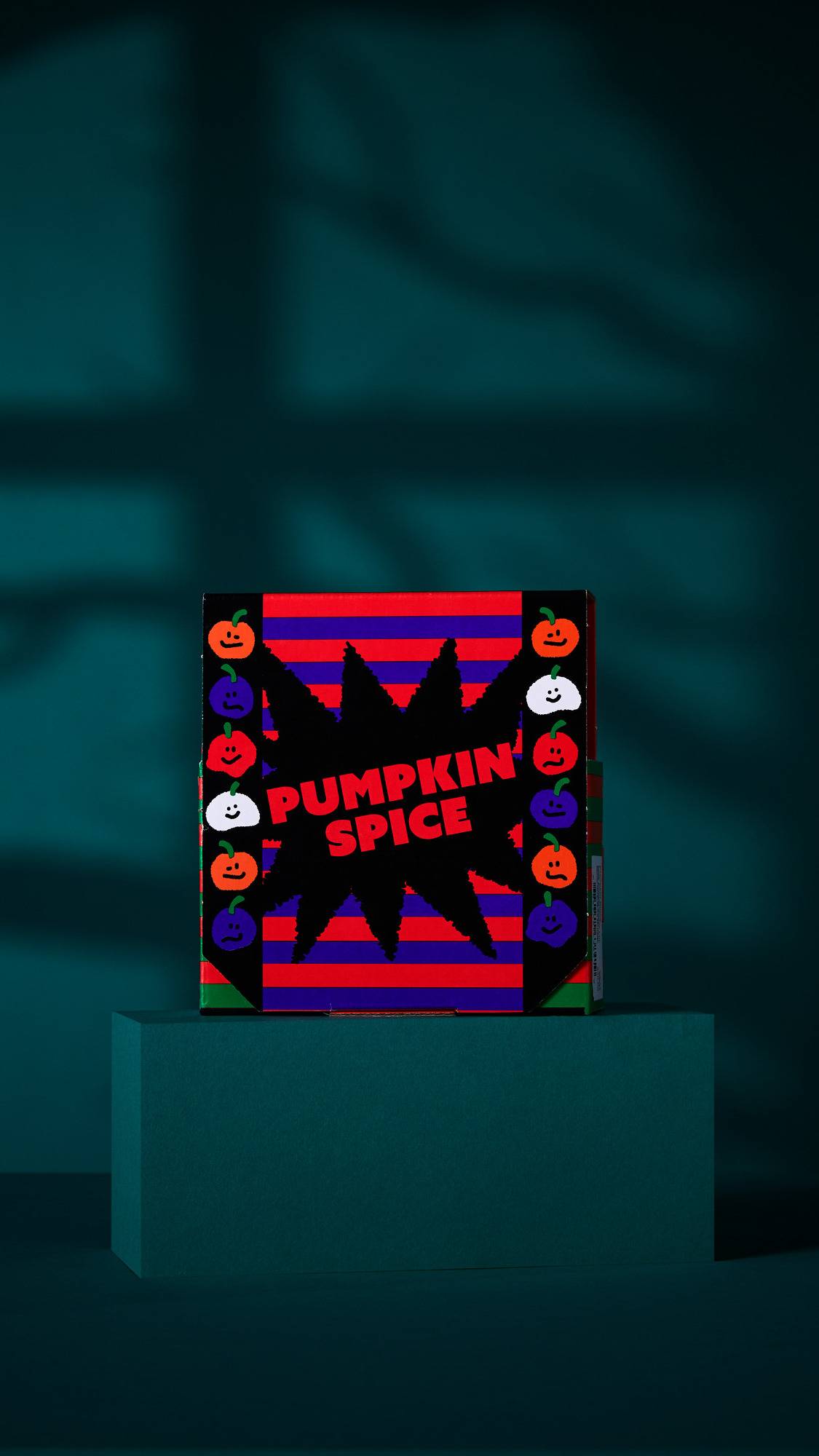The image shows a 'PUMPKIN SPICE' gift box. A striped pattern with a central starburst and pumpkin faces lines its border. The box sits on a dark teal platform, against a shadowed teal background.