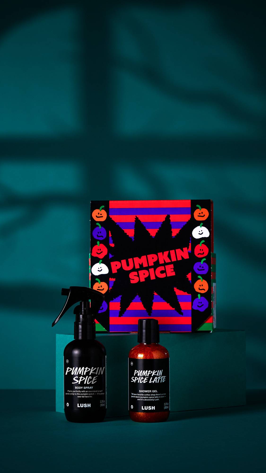 The image shows the 'Pumpkin Spice' gift set. A striped box with a starburst design sits on a dark teal platform behind a body spray and shower gel. The products and box are set against a dark teal background with shadows.