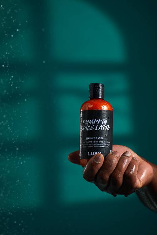 The image shows a dark-toned hand holding a bottle of 'Pumpkin Spice Latte' shower gel. Glittery orange liquid and a black label are visible, with water droplets on the hand and against a dark teal background.
