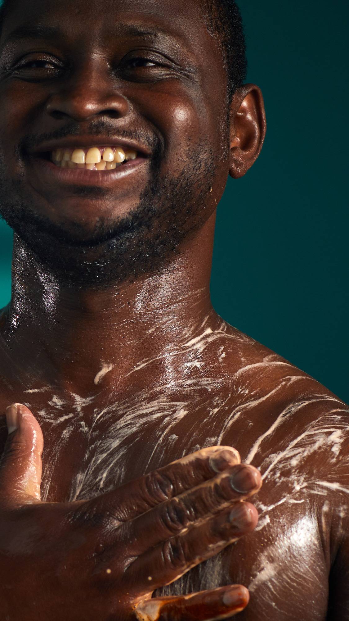 
The image shows a person with a joyful expression, lathering their chest and shoulder. A hand massages a thick, white, bubbly lather with an orange tint of the Pumpkin Spice Latte shower gel onto their skin, against a dark teal background.
