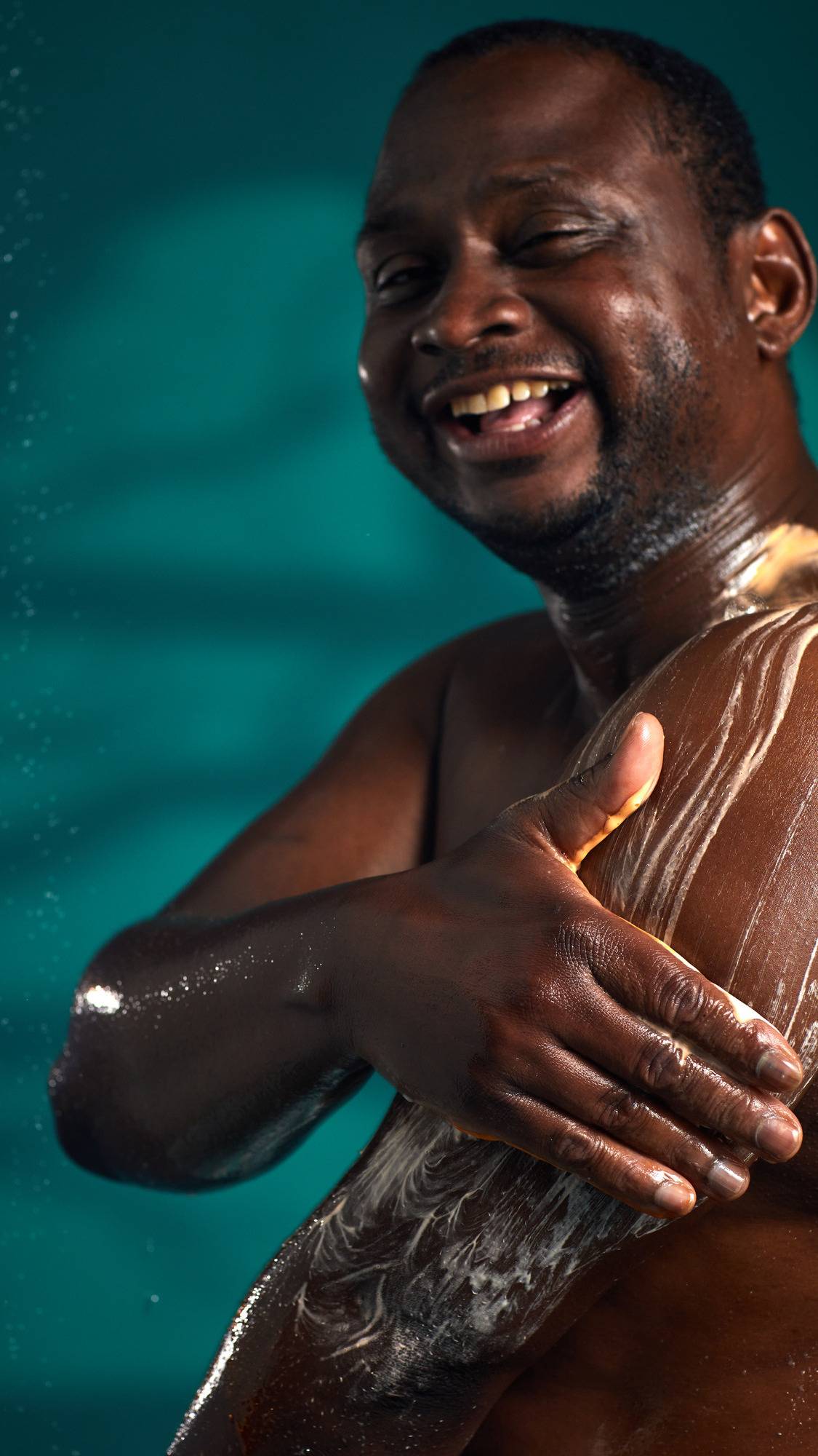 The image shows a person with a joyful expression, massaging a white, creamy lather onto their arm from the Pumpkin Spice Latte shower gel. A hand rubs the lather onto the skin, against a dark teal background.