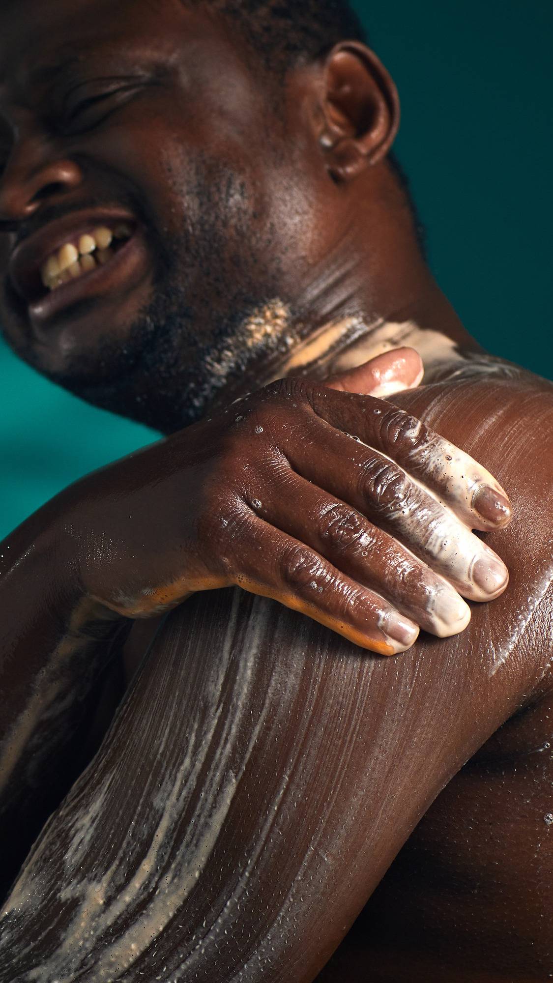 The image shows a person with a joyful expression, lathering their chest and shoulder. A hand massages a thick, white, bubbly lather with an orange tint of the Pumpkin Spice Latte shower gel onto their skin, against a dark teal background.