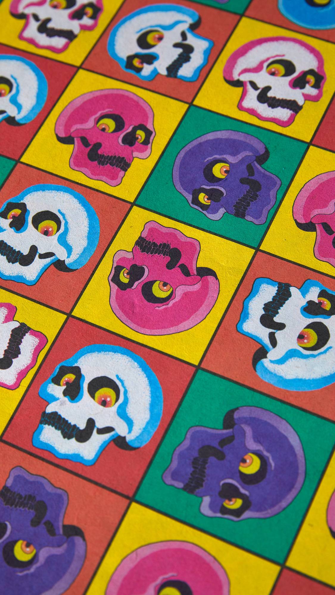 The image shows a close-up of the 'Skullywood' lokta paper. The multi-coloured grid pattern features cartoonish skulls in various colours, against a background of bright pink, red, green, and yellow squares.