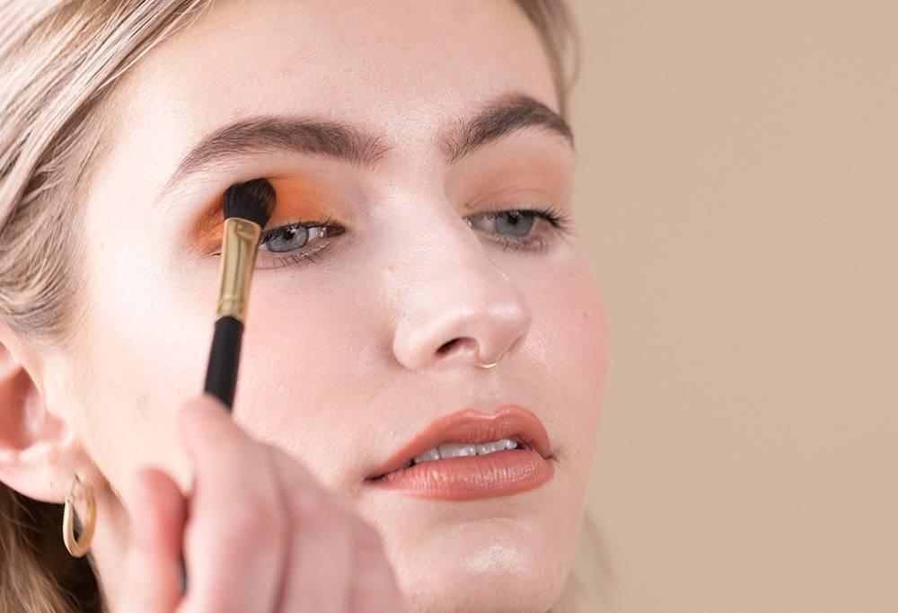 A person wearing peachy, orangey lipstick applies a soft orange eyeshadow using Hey Big Blender, a small blending brush.