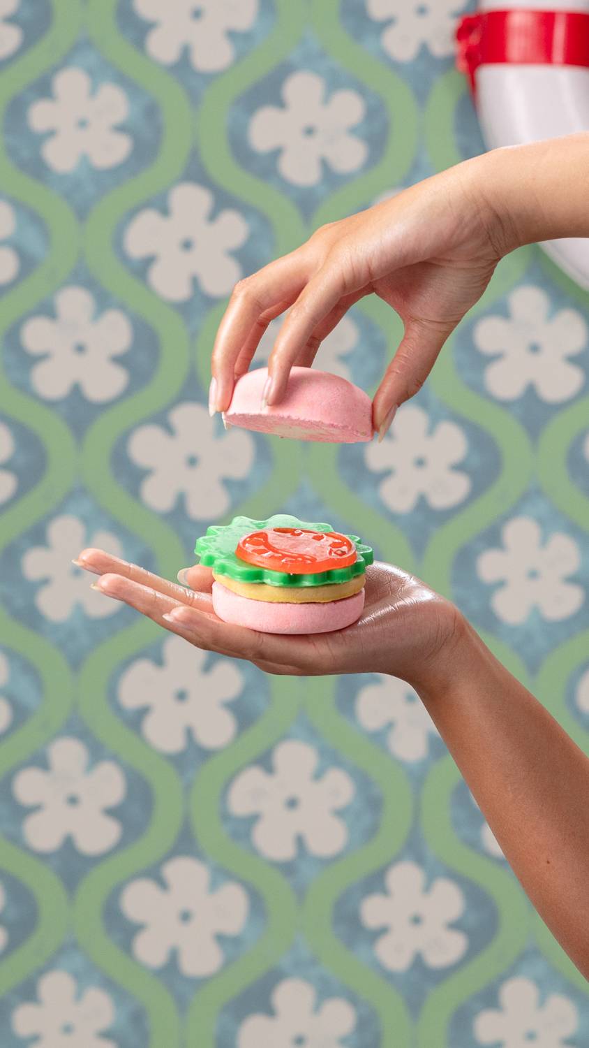 Model is holding the stacked burger-style Krabby Bathy gift in one hand, while the other is taking off the top bun bath bomb.
