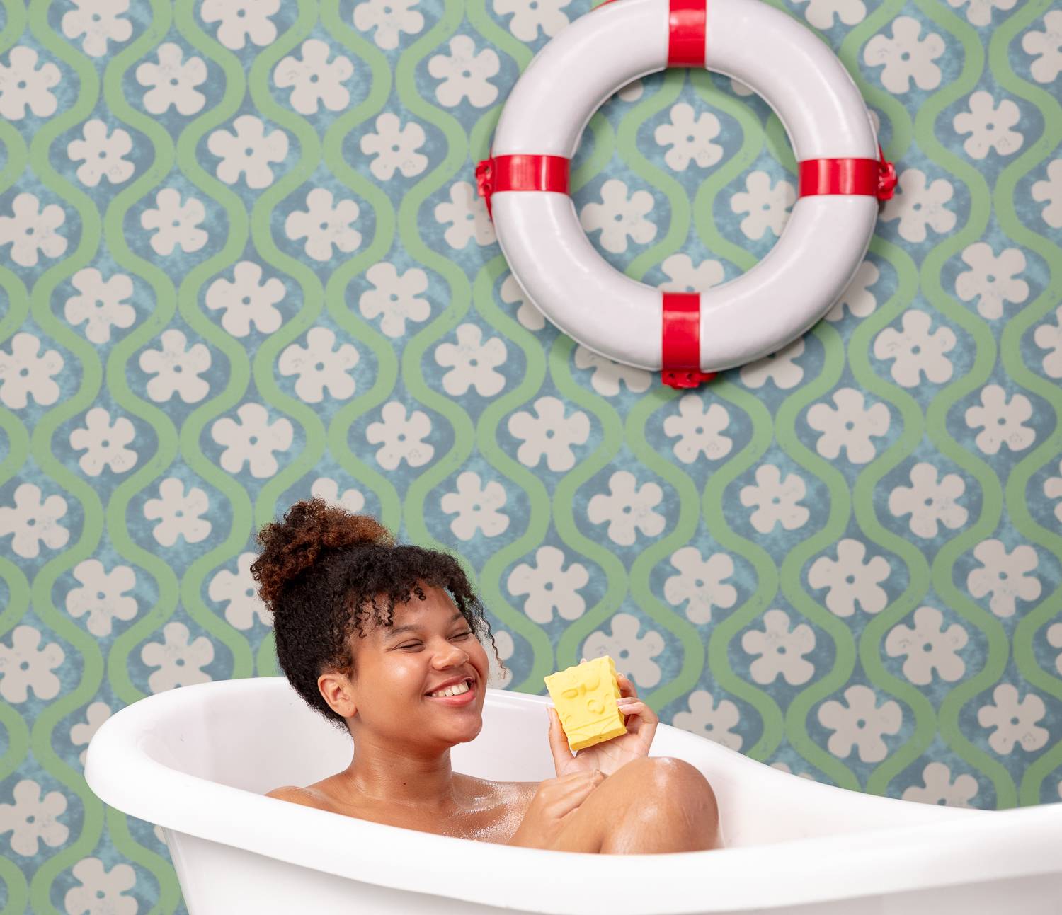 Model is in the bath in a fun, nautical-themed bathroom. They are smiling as they hold the product.