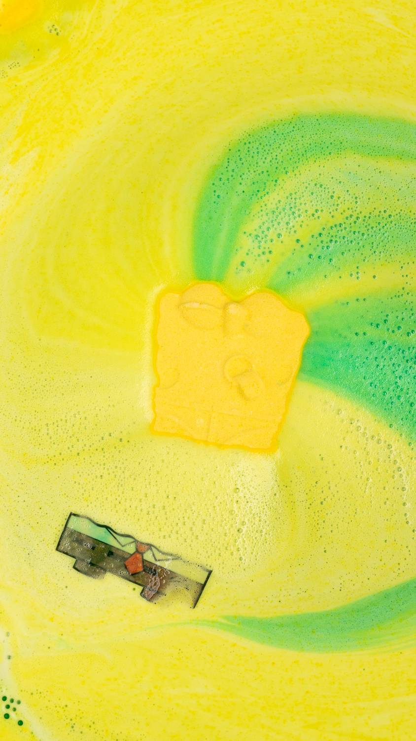 Spongebob bath bomb sits among bright swirls of green and yellow. Rice paper clothes can be seen floating.