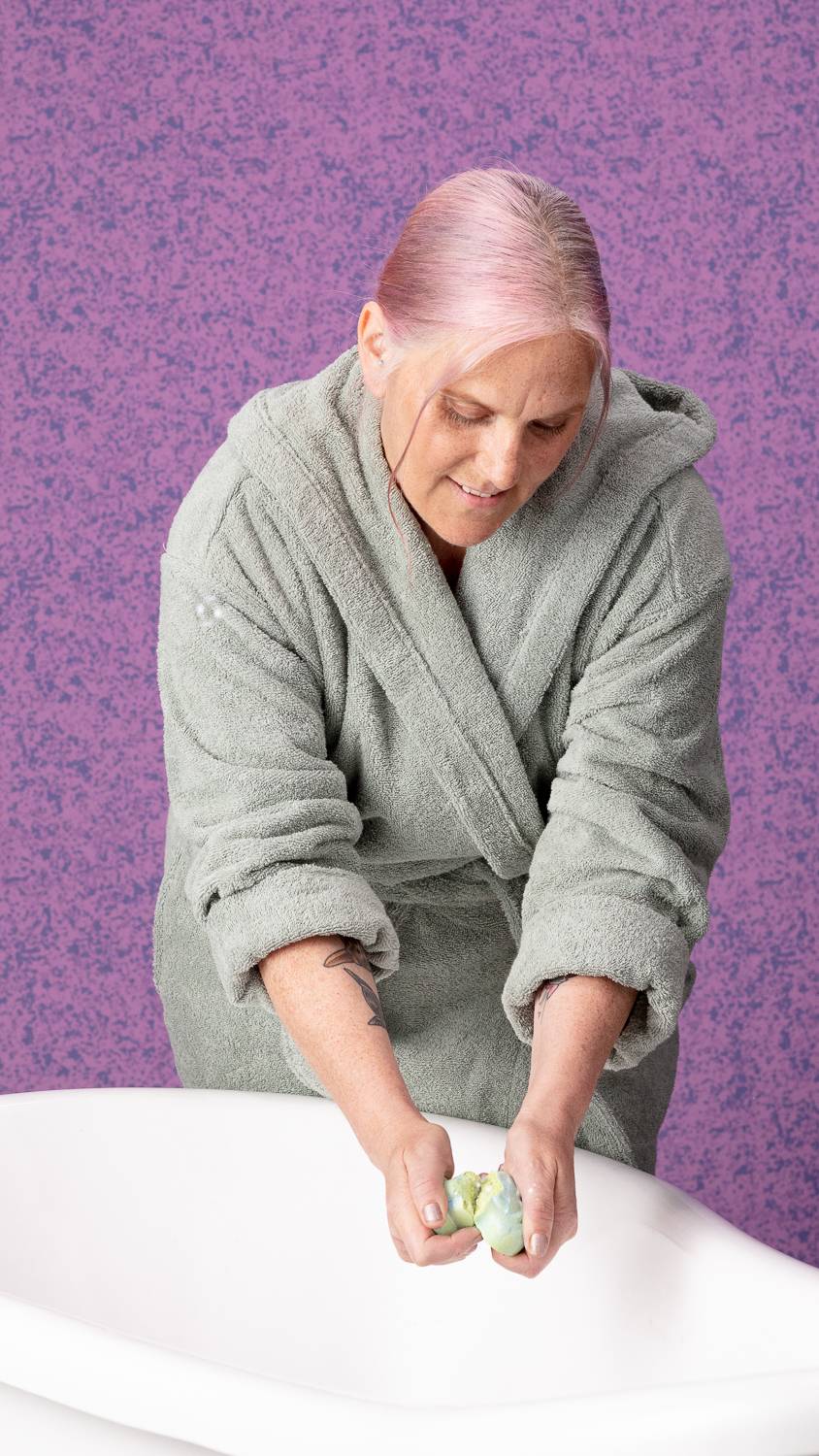 Model is stood over a bath crumbling the bubble bar in their hands. They wear a dressing gown with a purple-speckled background.