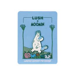 A 'Lush x Moomin' limited edition pin badge on a light blue backing card. The enamel pin depicts Moomintroll walking through colourful flowers with blue soap suds on his nose, head, and tail.