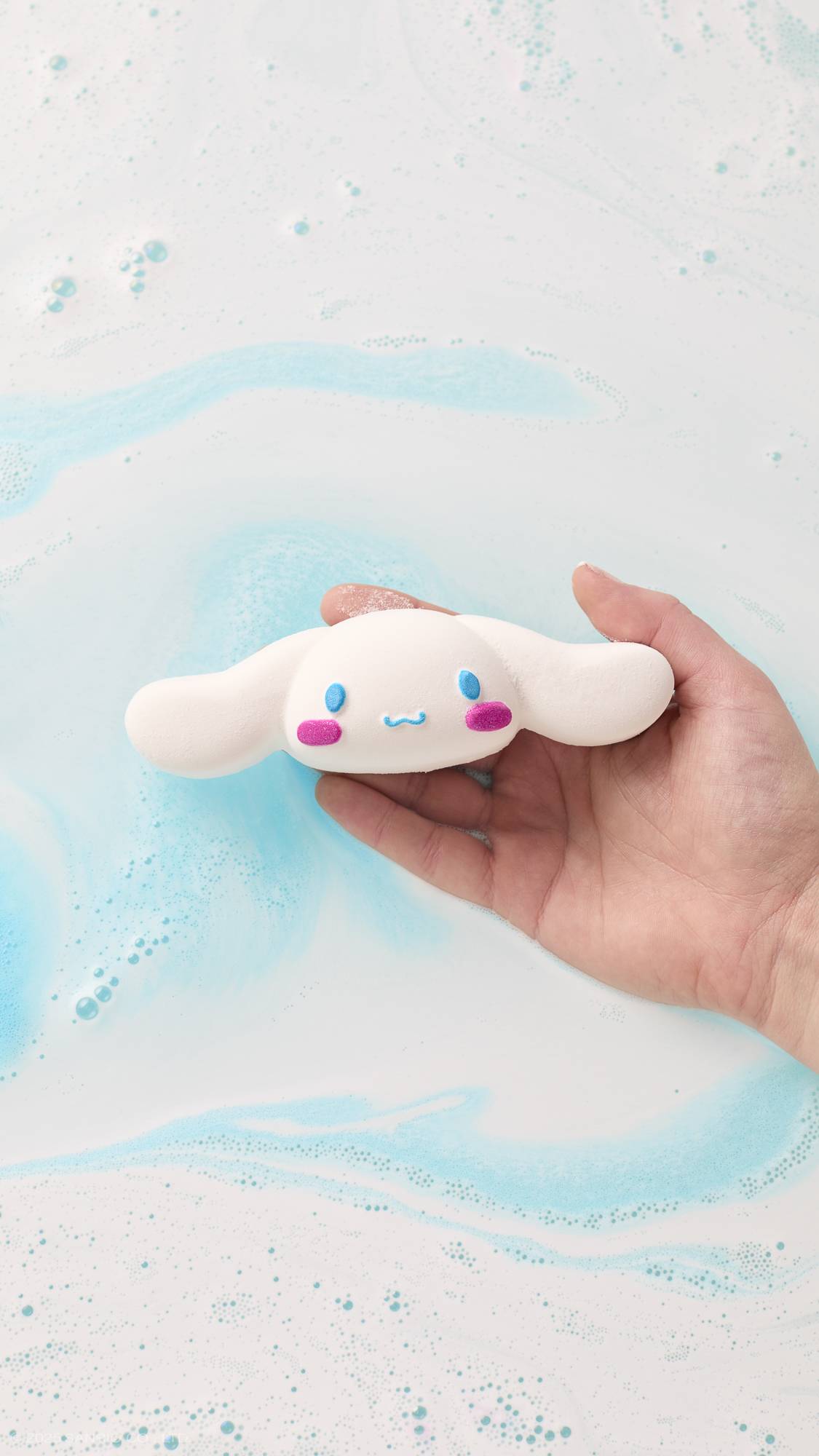 An aerial view image of the model's hand reaching in from the right-hand side, holding the white, long-eared Cinnamoroll bath bomb in their palm above a sea of velvety, white foam over glacier blue water below.