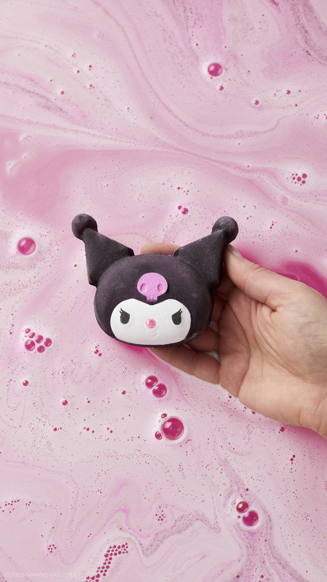 An aerial view image of the model's hand reaching in from the right-hand side, holding the white, pink and black Kuromi bath bomb in their palm above a sea of velvety, pastel pink foam over vibrant pink water below.