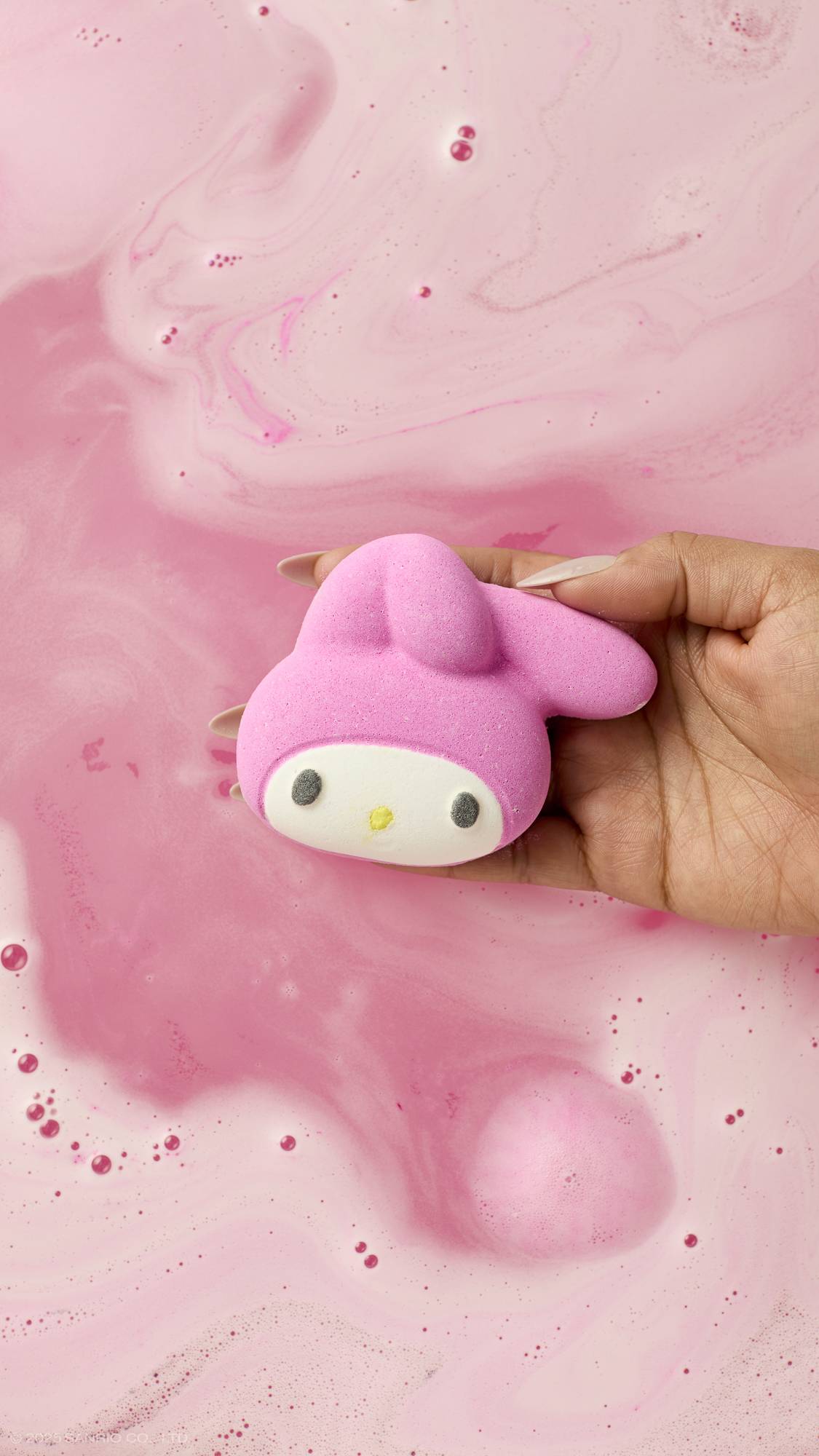 An aerial view image of the model's hand reaching in from the right-hand side holding the white and pink, rabbit-faced My Melody bath bomb in their palm above pastel pink bath water below. 