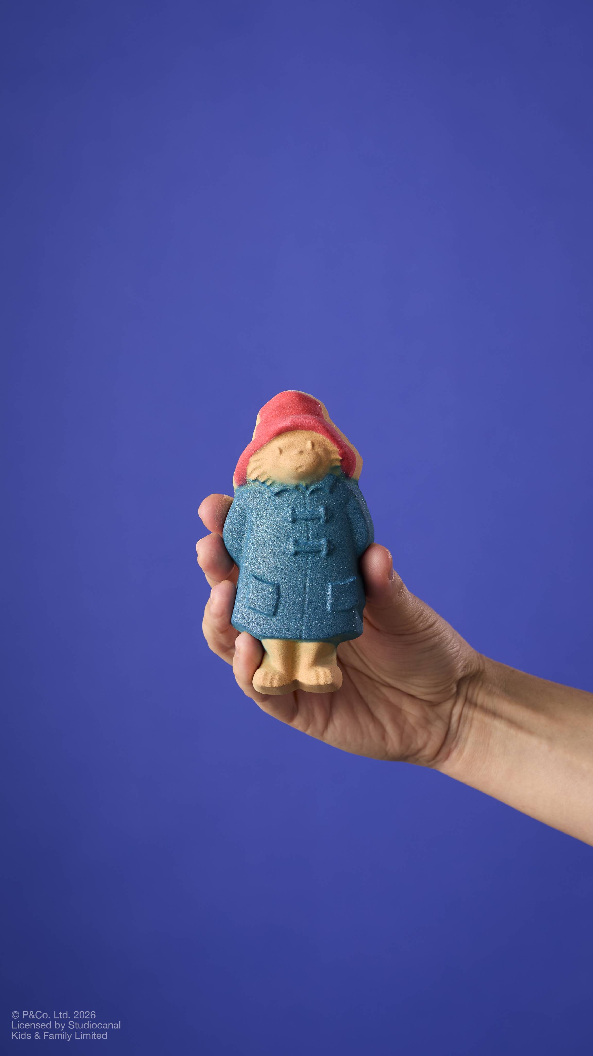 A model's arm holds a "Paddington Bear" bath bomb against a solid blue background. The bear wears his signature red hat and blue duffle coat with textured button details. 