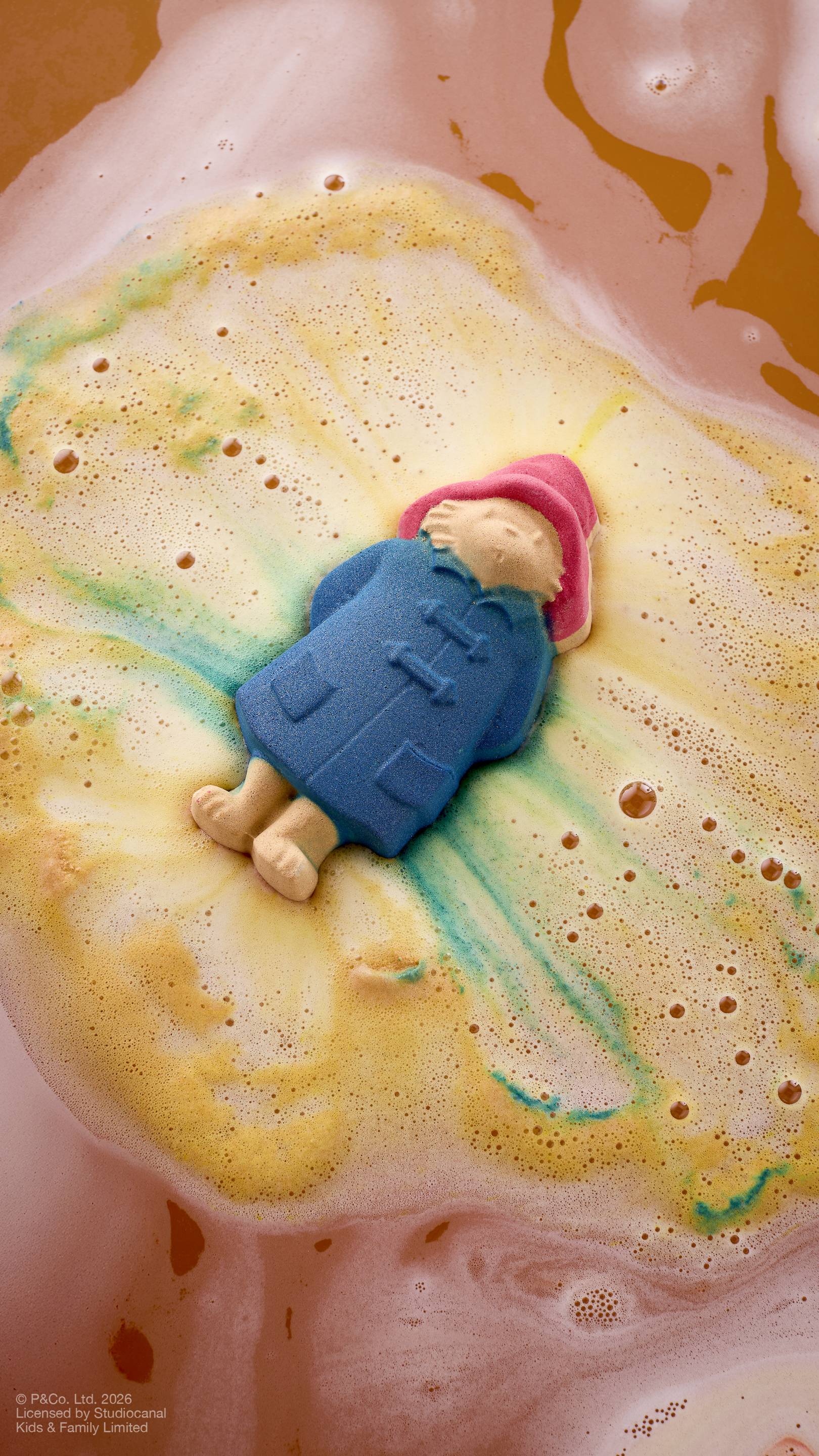 An aerial view image of the "Paddington Bear" bath bomb floating on the surface of bath water, creating a swirl of yellow, orange, and green foam. The bear, in his red hat and blue coat, is partially submerged as the product dissolves. 