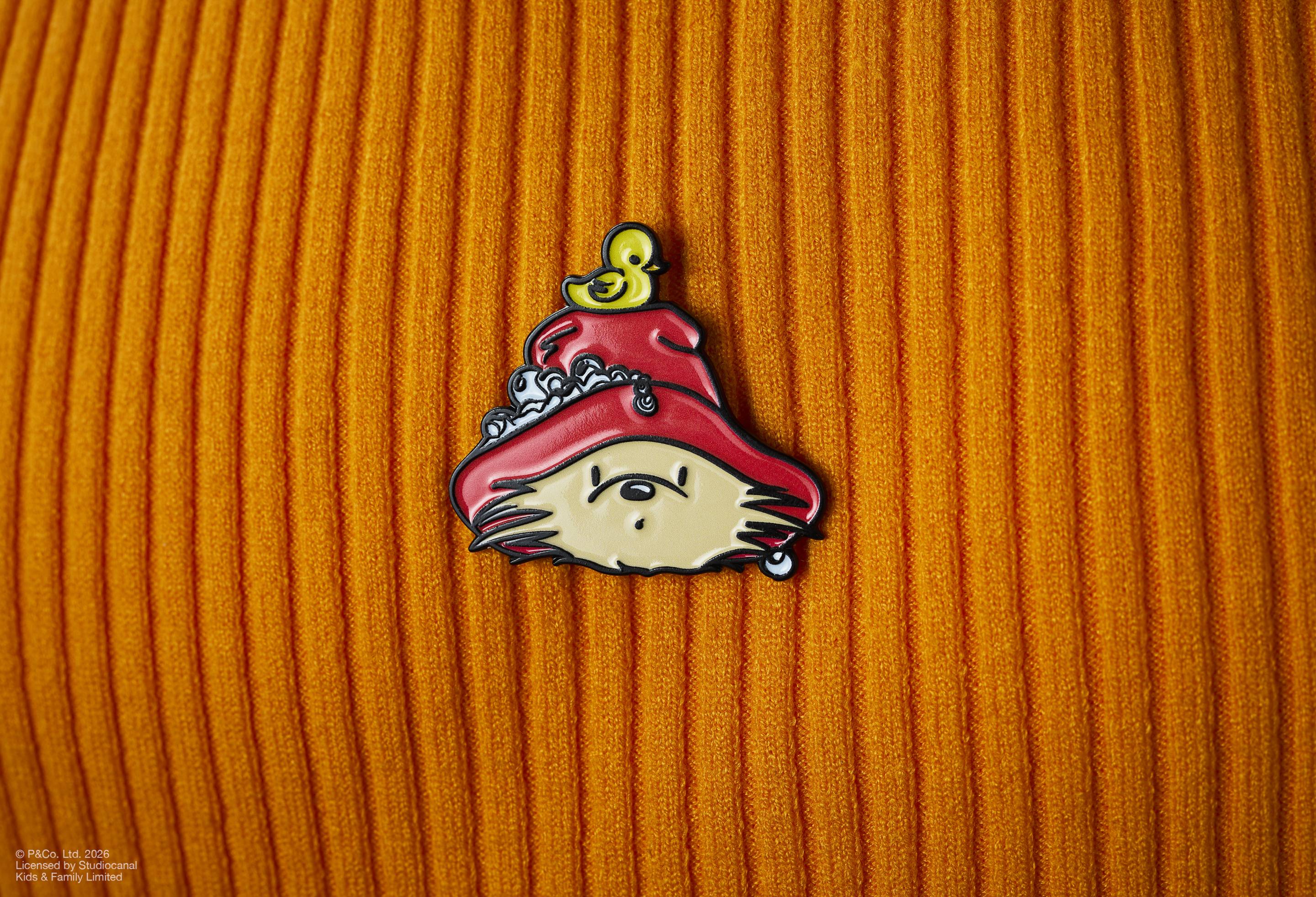 The image shows a Paddington x Lush enamel pin badge pinned to an orange ribbed, corduroy fabric. The badge features Paddington Bear’s head in a red hat with a yellow rubber duck on top and soap bubbles.