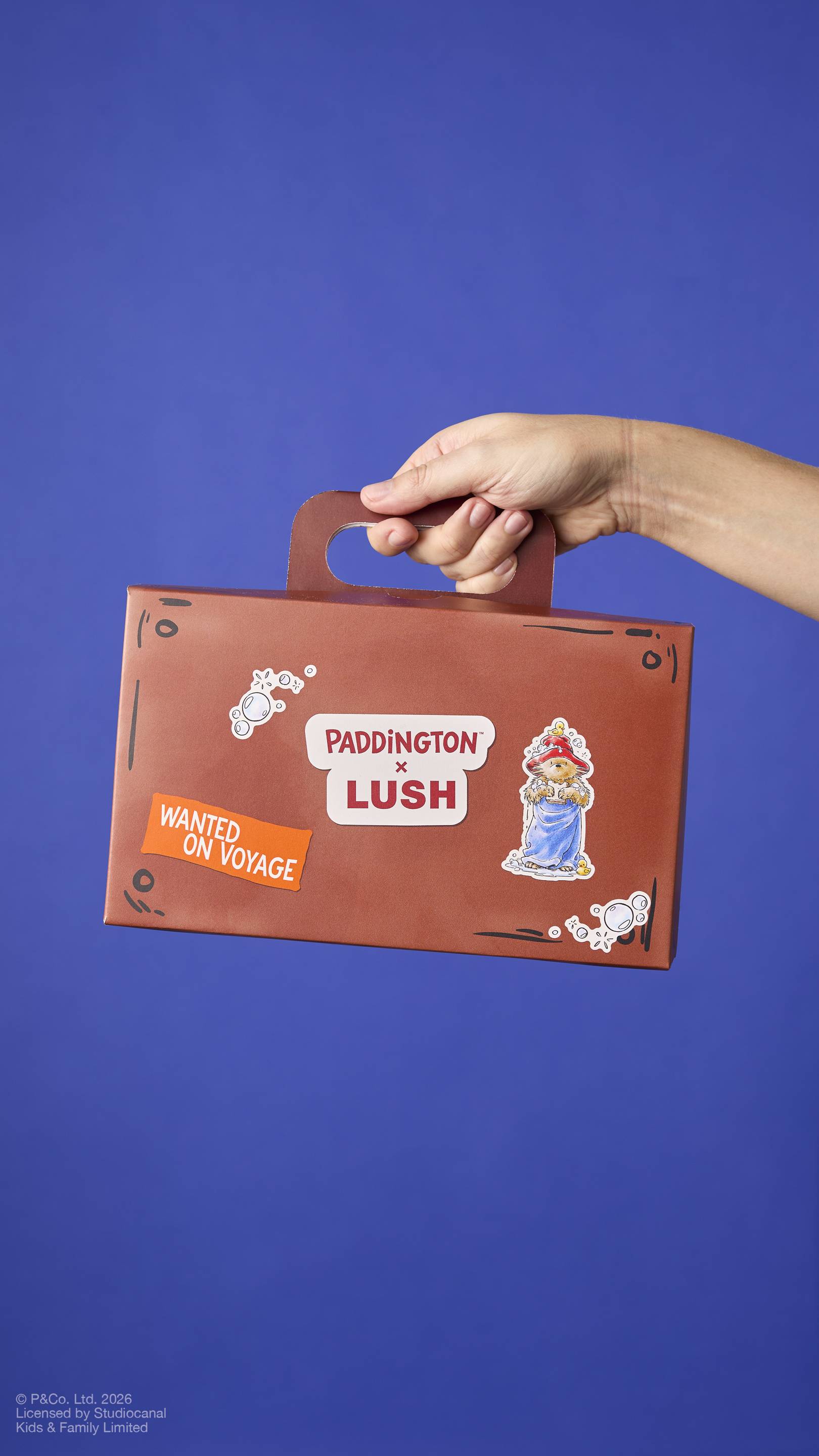 A brown cardboard gift box designed to look like a small suitcase, held by hand by its handle. It features ‘PADDINGTON x LUSH’ branding, an illustration of the bear in a towel, and a ‘WANTED ON VOYAGE’ sticker against a blue background.