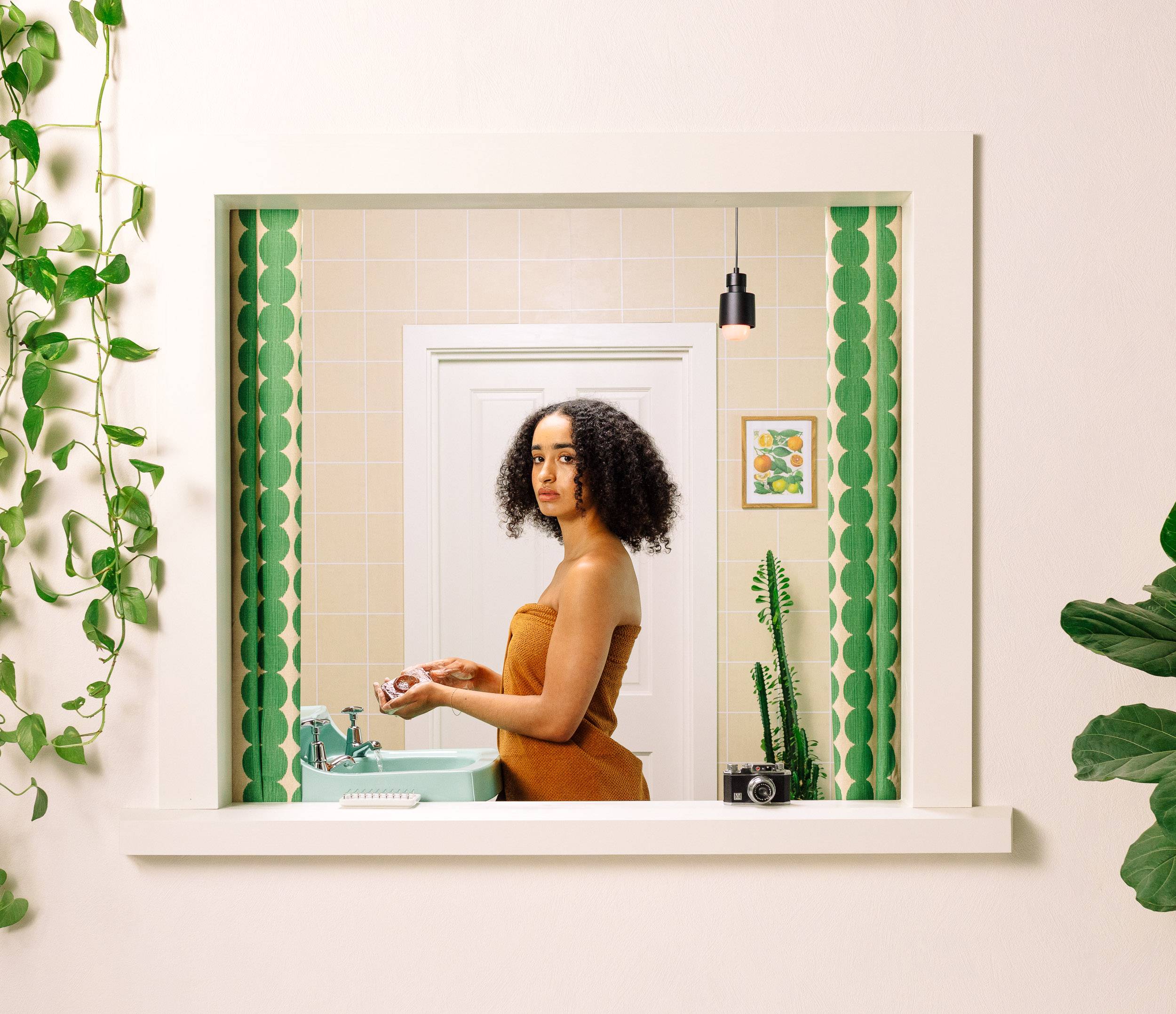 Model stands in a retro-styled bathroom in a sand-coloured towel over a teal sink holding the soap.