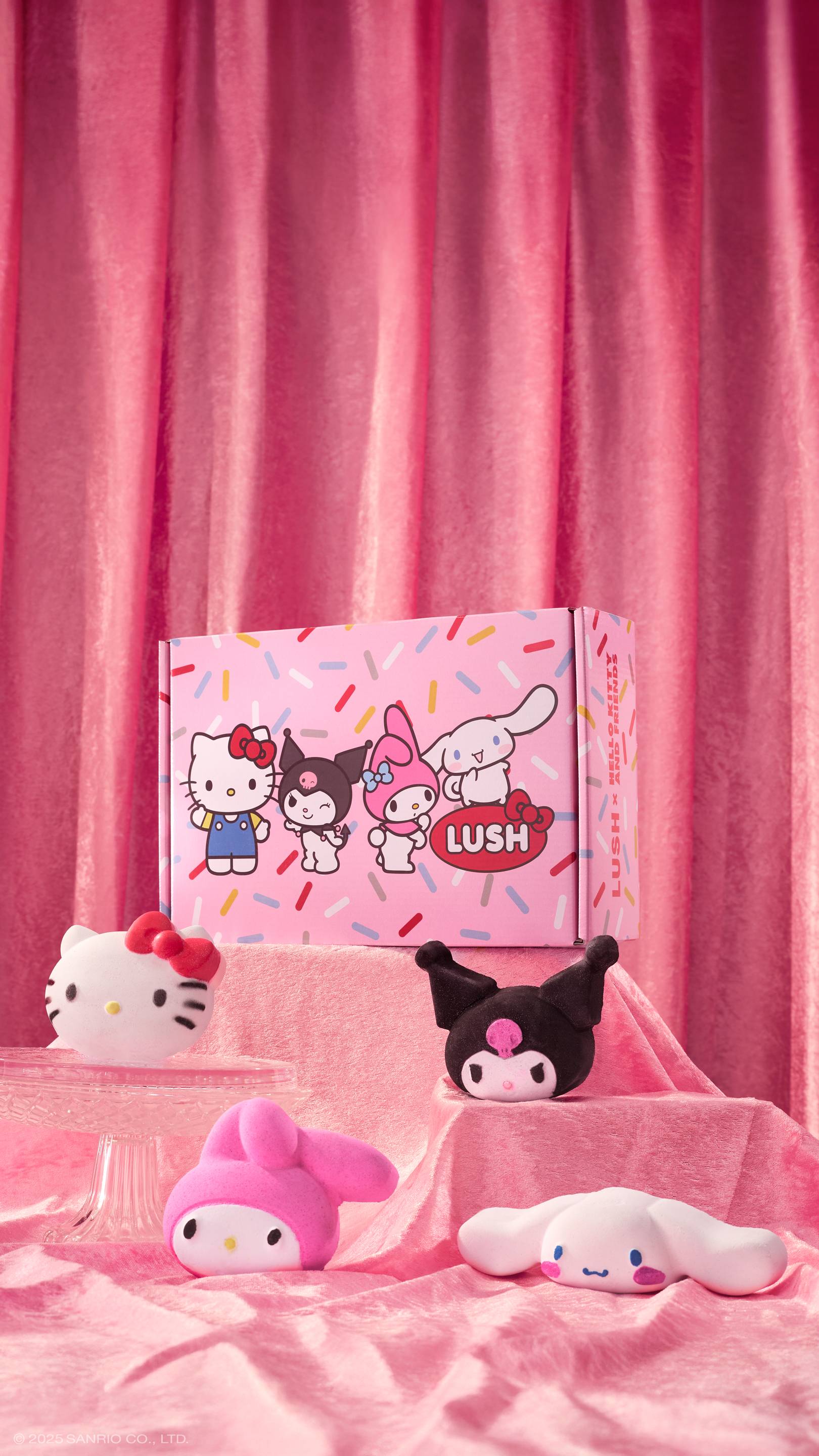 The Hello Kitty and Friends gift set and bath bombs are displayed on pink velvet fabric with ruffled folds. The box with cartoon illustrations sits higher up, surrounded by four bath bombs shaped like Hello Kitty, My Melody, Kuromi, and Cinnamoroll.