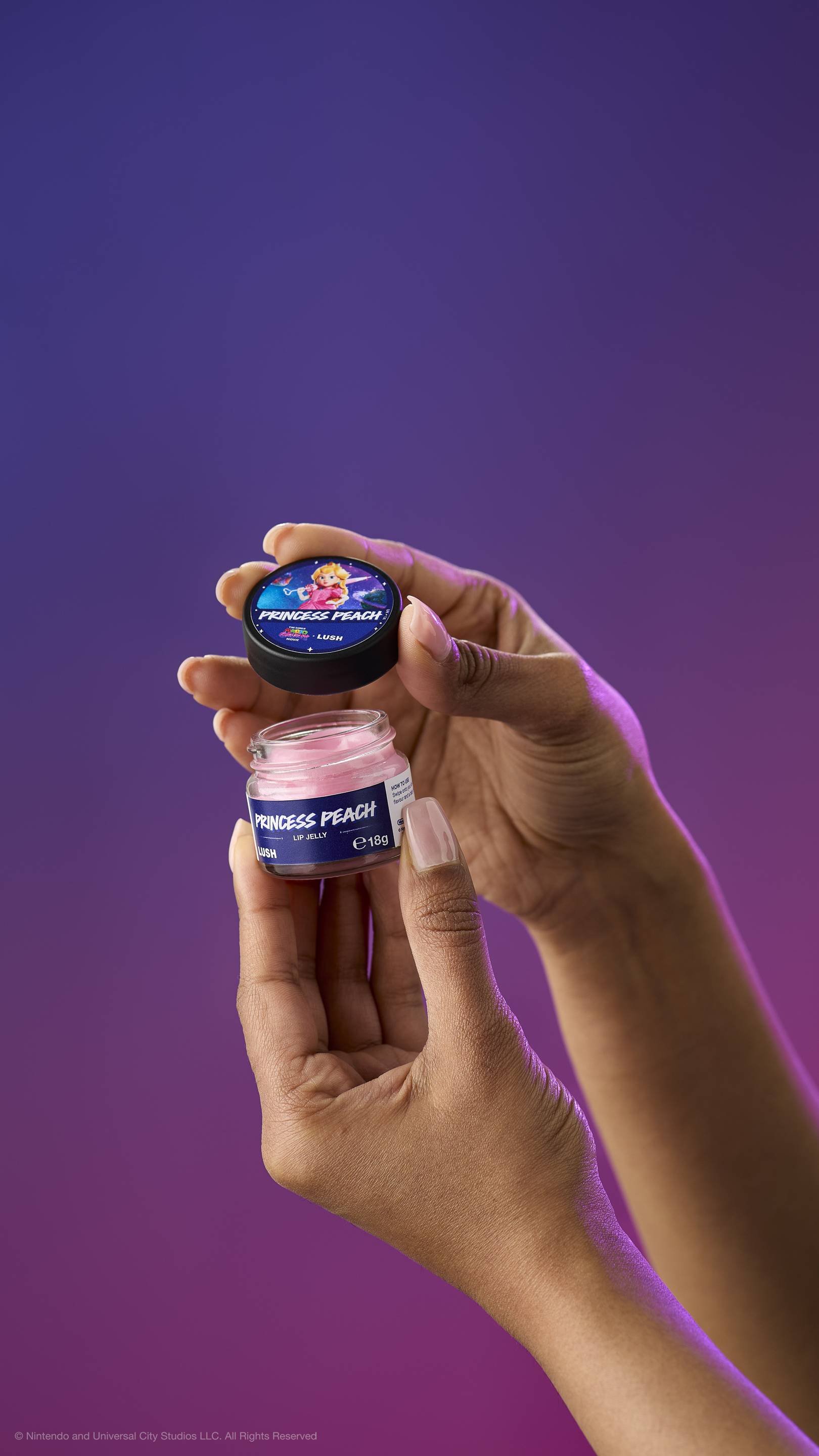 Two hands hold an open pot of creamy pink Princess Peach lip jelly and its lid against a purple-magenta gradient. The lid features Princess Peach holding a heart-shaped wand.