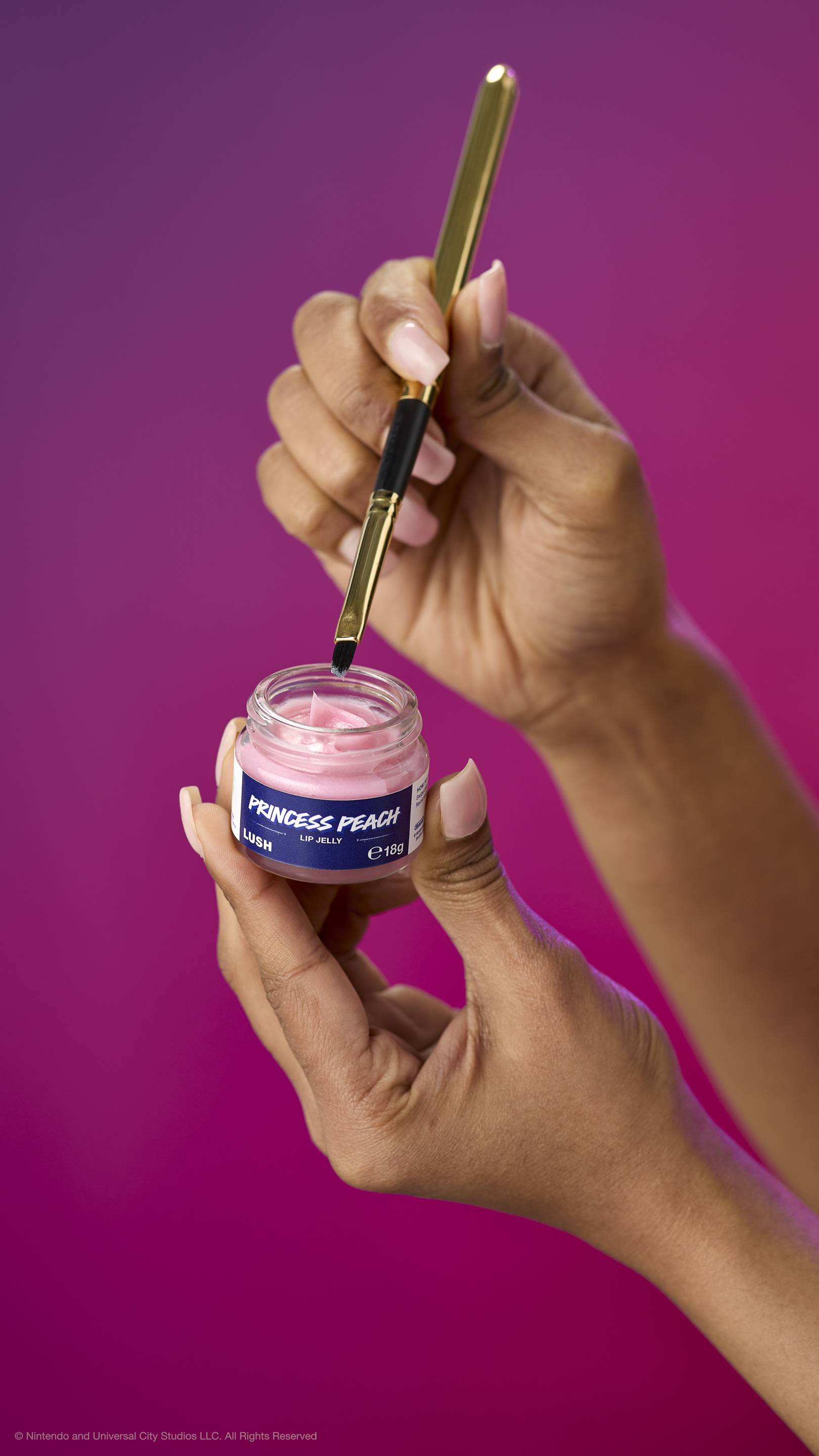 Hands hold an open pot of creamy pink Princess Peach lip jelly while a gold lip brush picks up product. Against a purple-magenta gradient background.