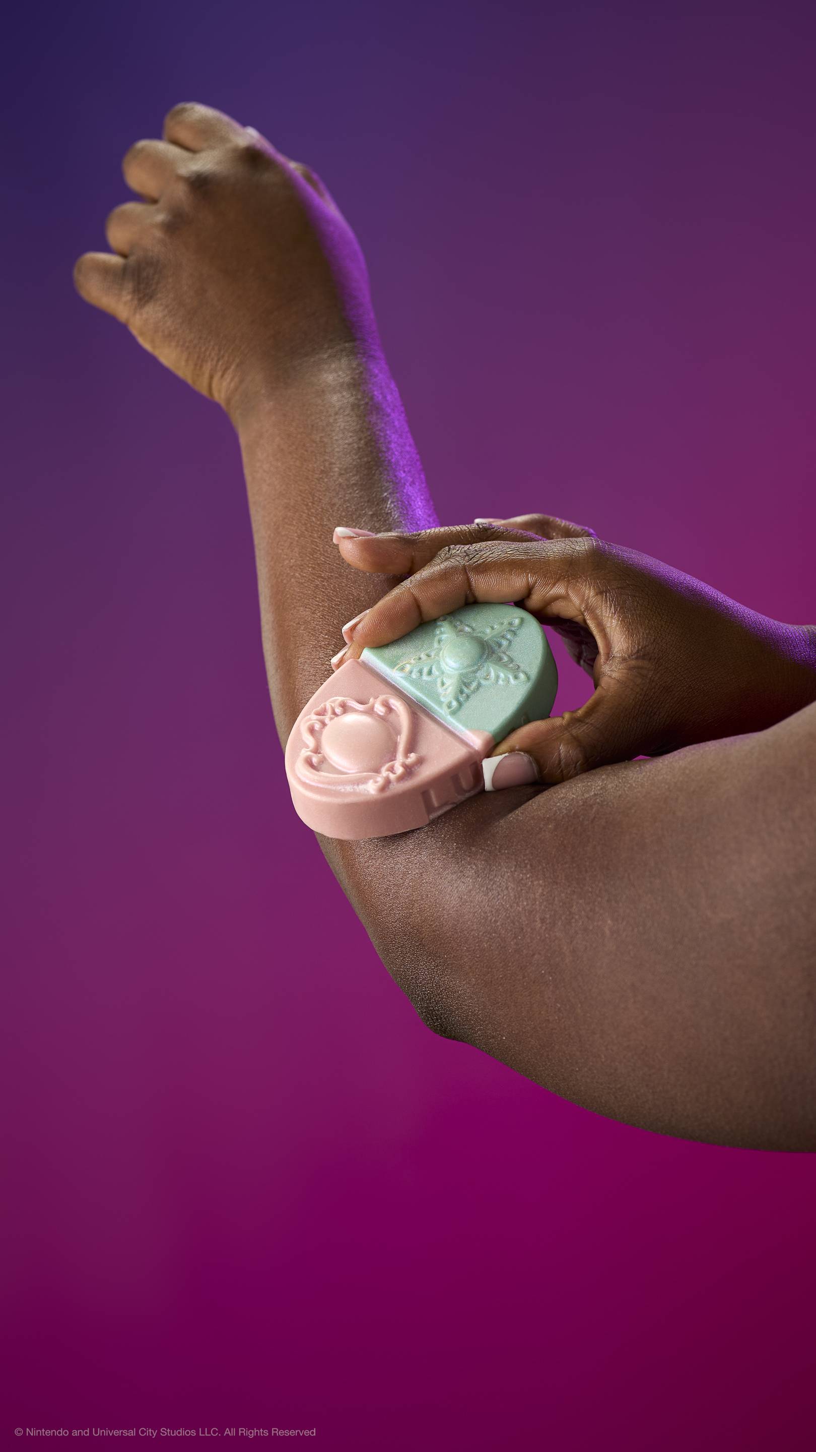 A hand glides the pink and teal Princess Power body balm over an elbow. Features embossed brooch and star designs. Against a purple-magenta gradient with plastic-free glitter.
