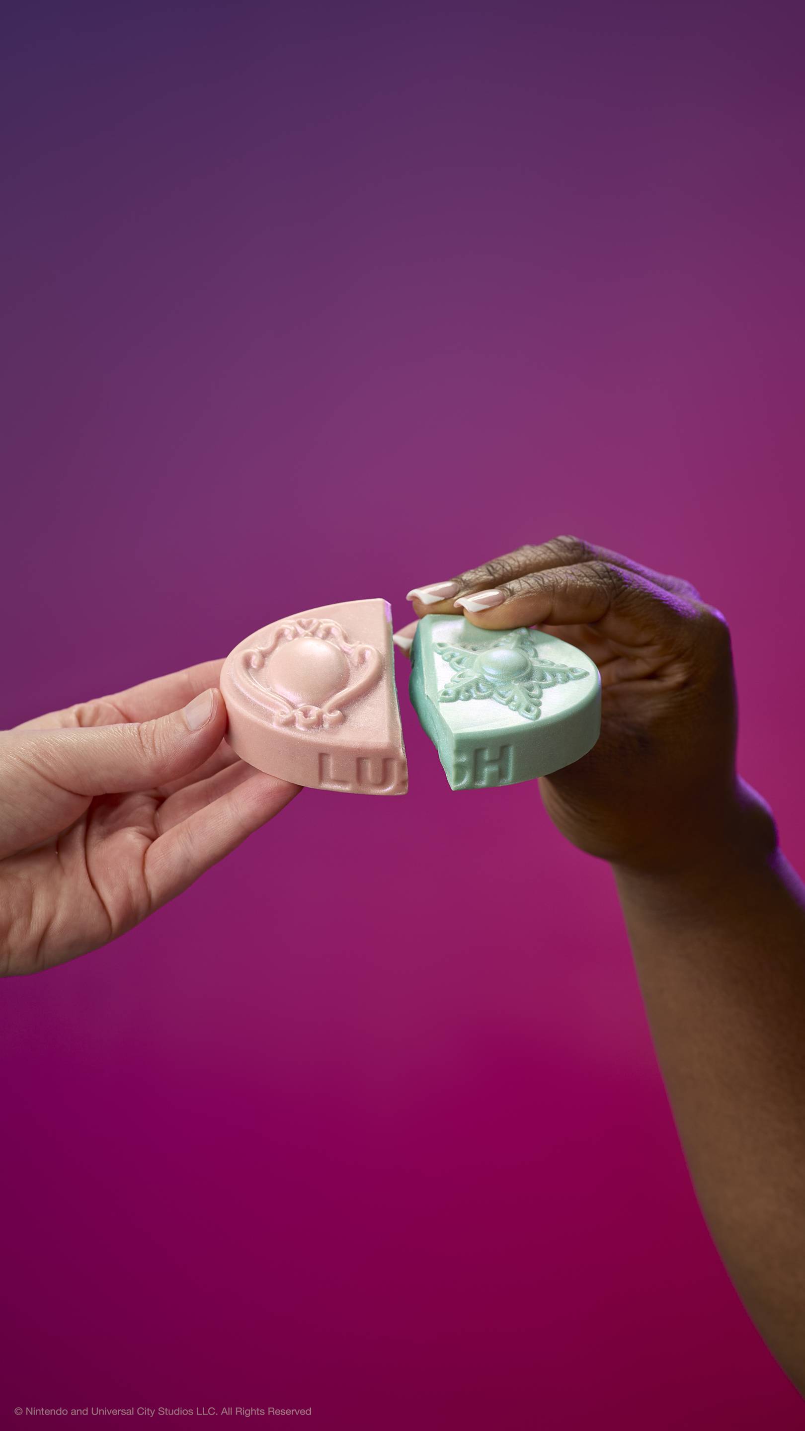 Two hands join the pink and teal halves of the Princess Power body balm against a purple-pink gradient. The pieces feature embossed brooch and star designs and the LUSH logo on the side.