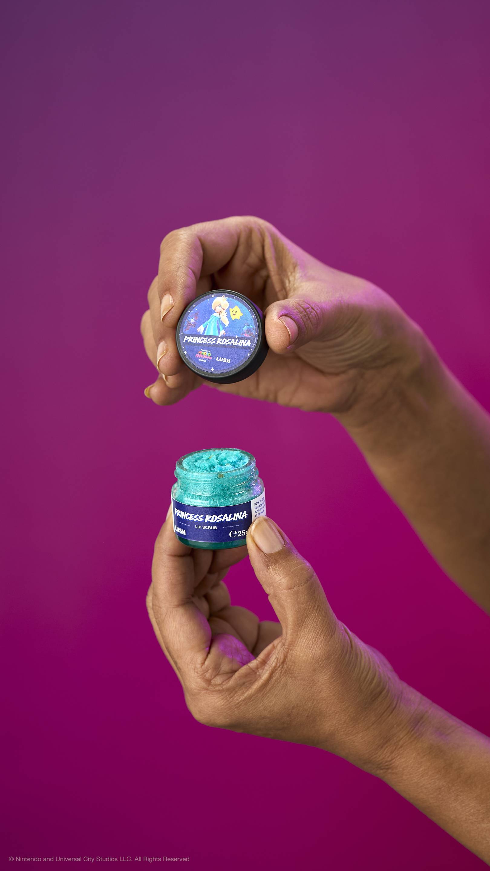 Two hands hold an open pot of turquoise Princess Rosalina lip scrub and its lid against a purple background. The lid features Princess Rosalina and a Luma character.