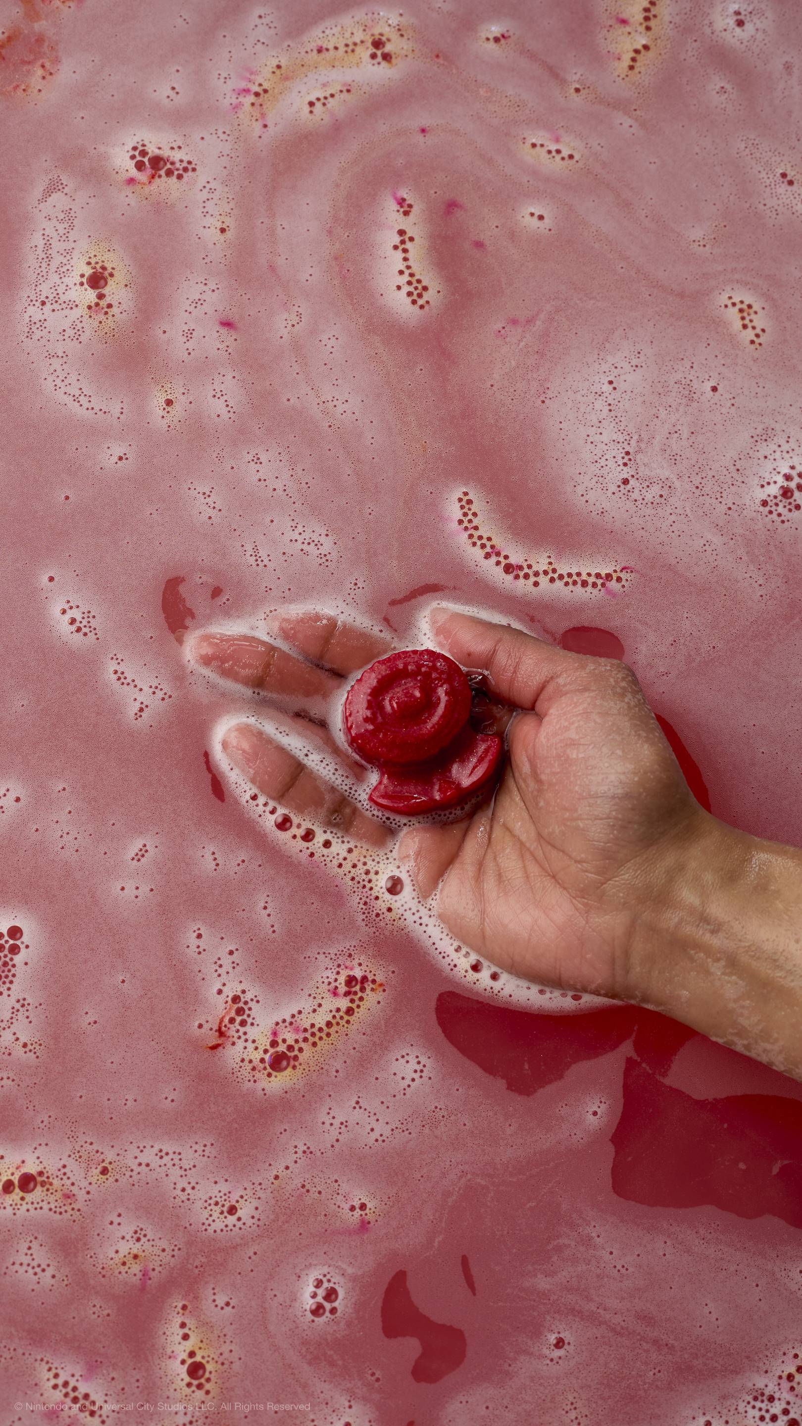 A hand holds a small, red soap shaped like a Fire Flower over bubbly, pink bathwater. This is the surprise insert found inside a Yoshi egg bath bomb. All glitter used is plastic-free.
