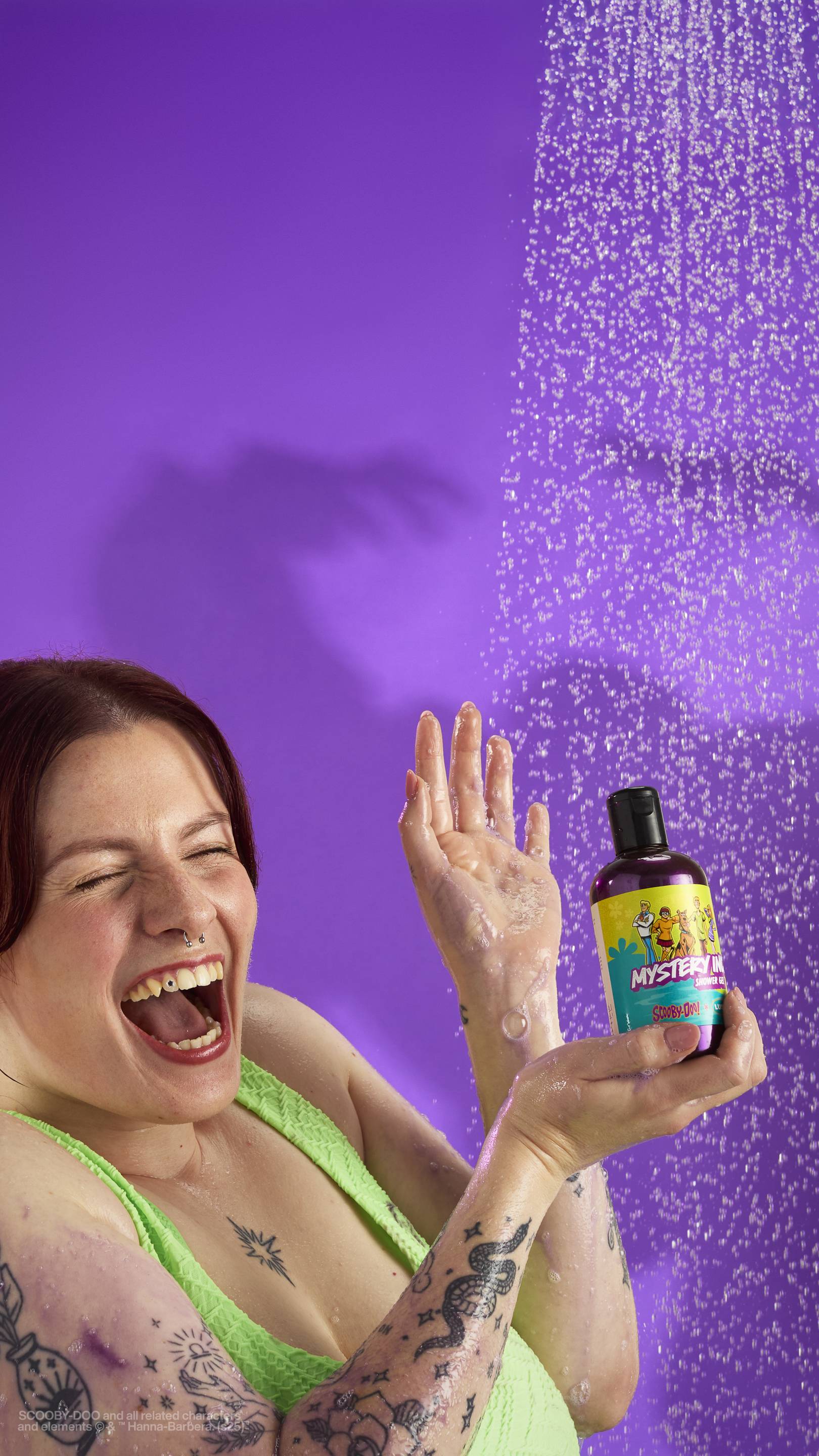 The image shows a solid purple background with out-of-focus falling shower water. A model is in a neon green bathing suit holding a clear plastic bottle of the shimmery, purple Mystery Inc. shower gel with a colourful label as droplets splash off. 