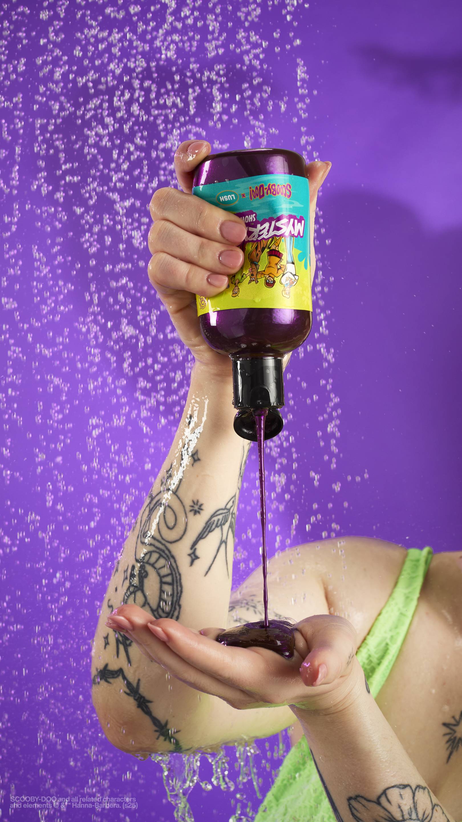 The image shows a solid purple background with out-of-focus falling shower water. A light-toned hand holds an upside-down clear plastic bottle of the shimmery, purple Mystery Inc. shower gel as the product pours into a cupped hand, held lower. 