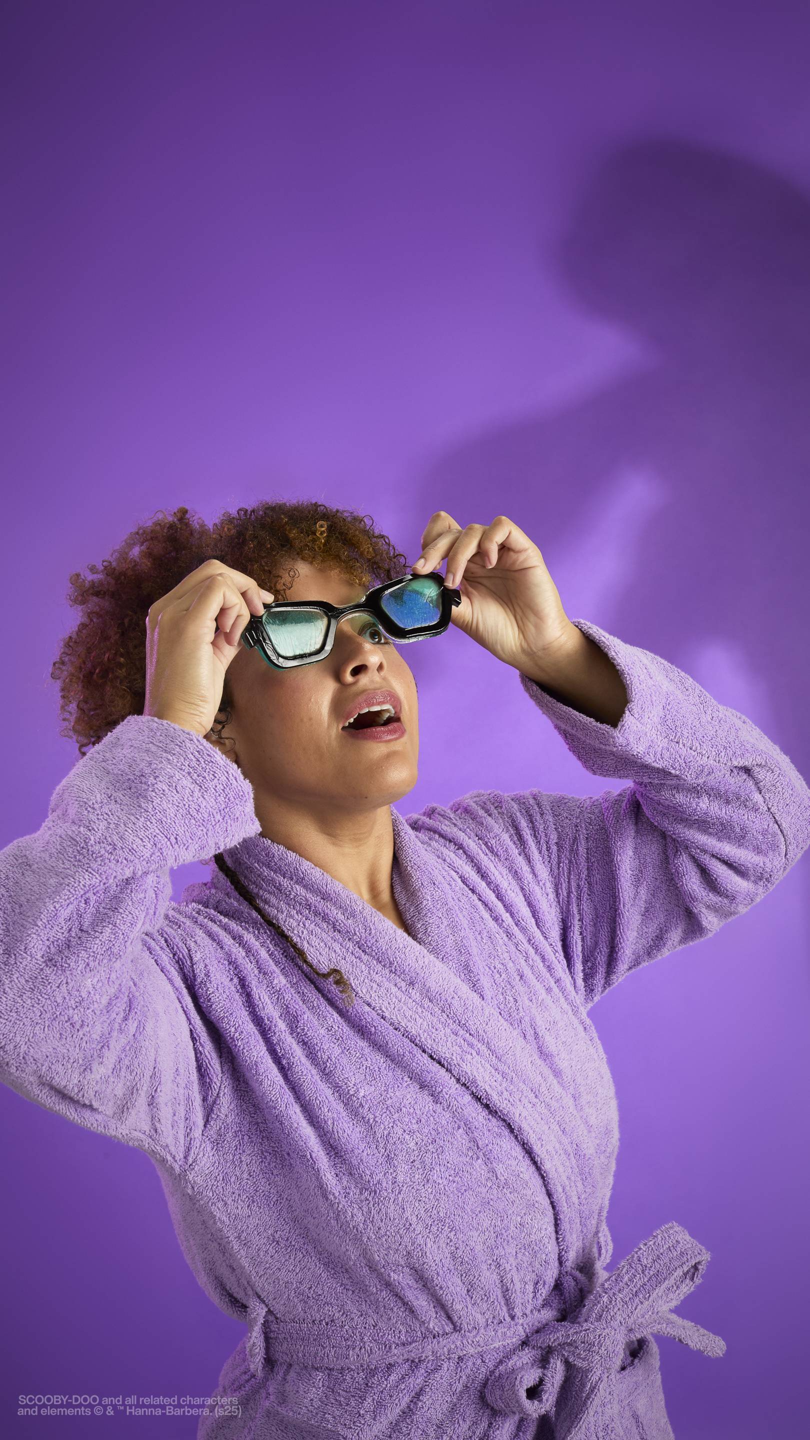The image shows a dark purple background with an eerie shadow just visible. A model with curly brown hair, wearing a purple robe, holds the My Glasses! jelly eye mask over their face as they make a scared face. 