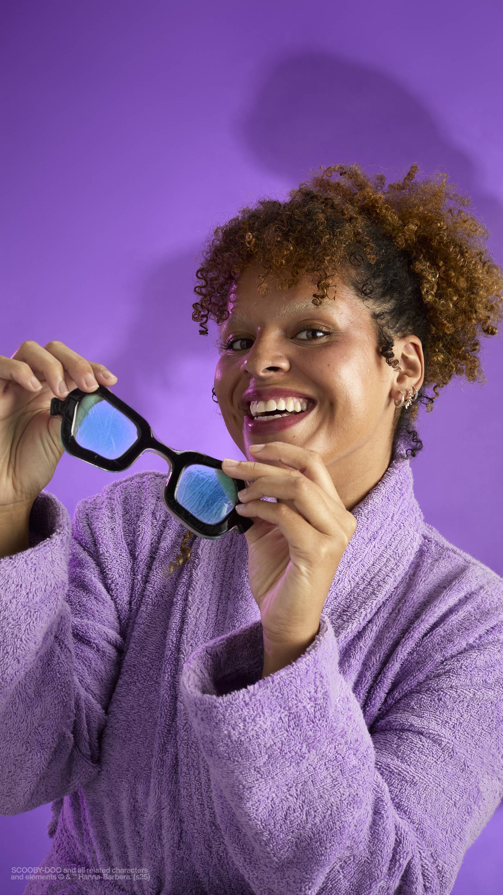 The image shows a dark purple background with an eerie shadow just visible. A model with curly brown hair, wearing a purple robe, holds the My Glasses! jelly eye mask in front of them as they smile widely.