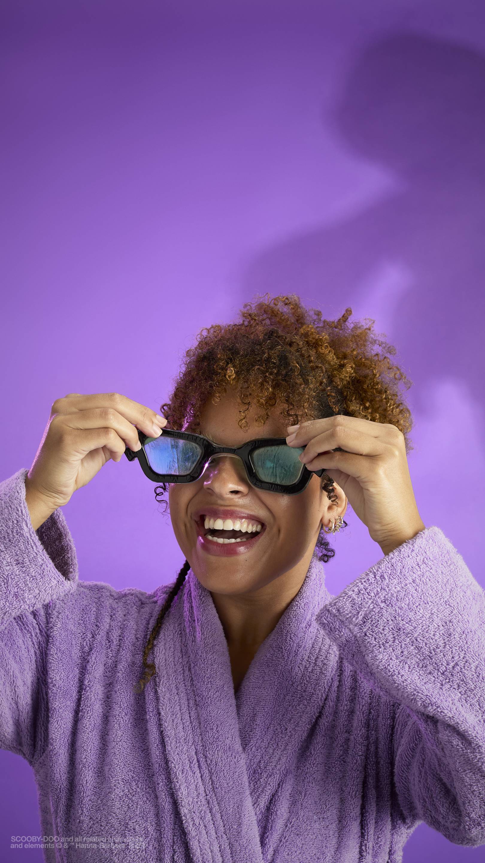 The image shows a dark purple background with an eerie shadow just visible. A model with curly brown hair, wearing a purple robe, holds the My Glasses! jelly eye mask in front of them as they smile widely.