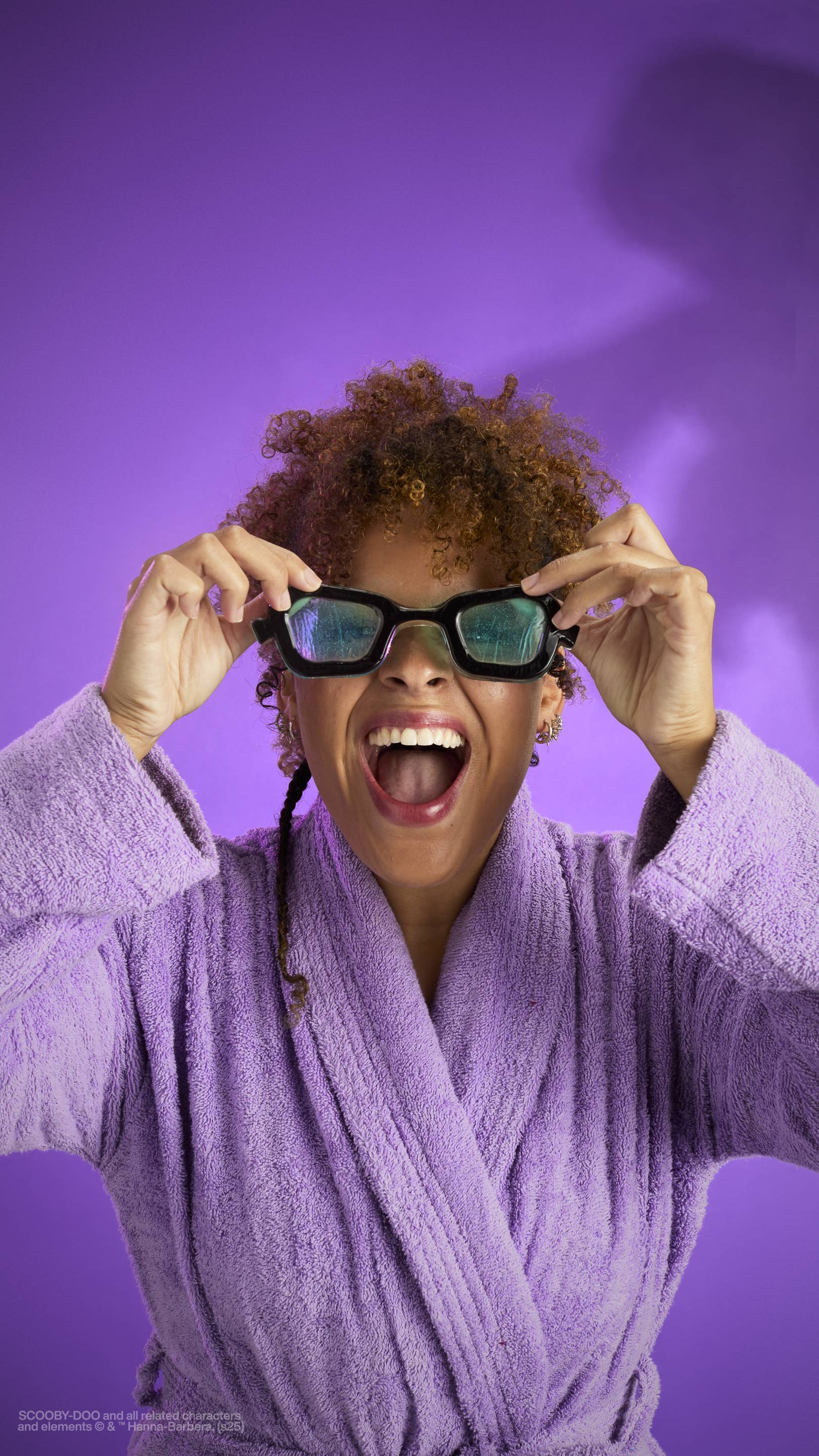 The image shows a dark purple background with an eerie shadow just visible. A model with curly brown hair, wearing a purple robe, holds the My Glasses! jelly eye mask over their face as they make a scared face. 