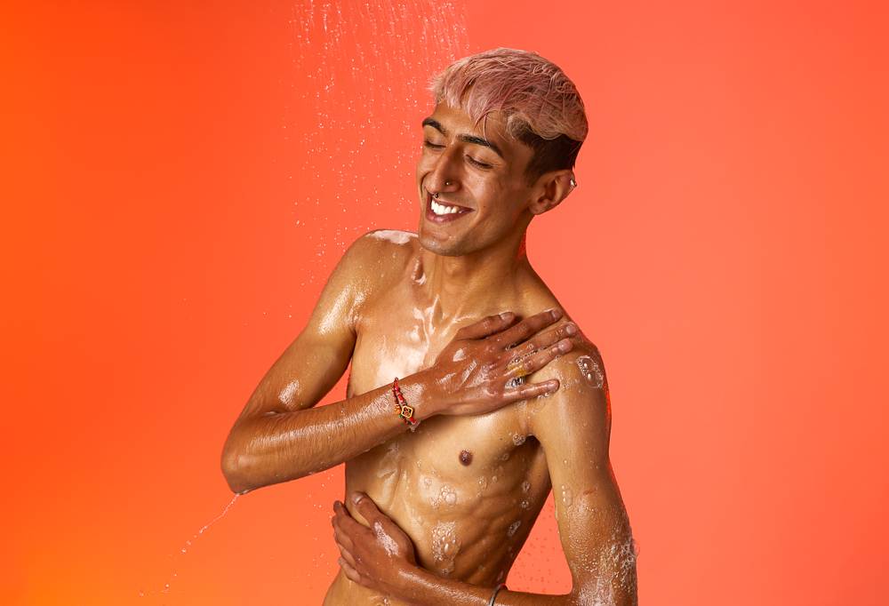 A smiling model lathers Mango shower gel on their body under a shower in front of an orange background.