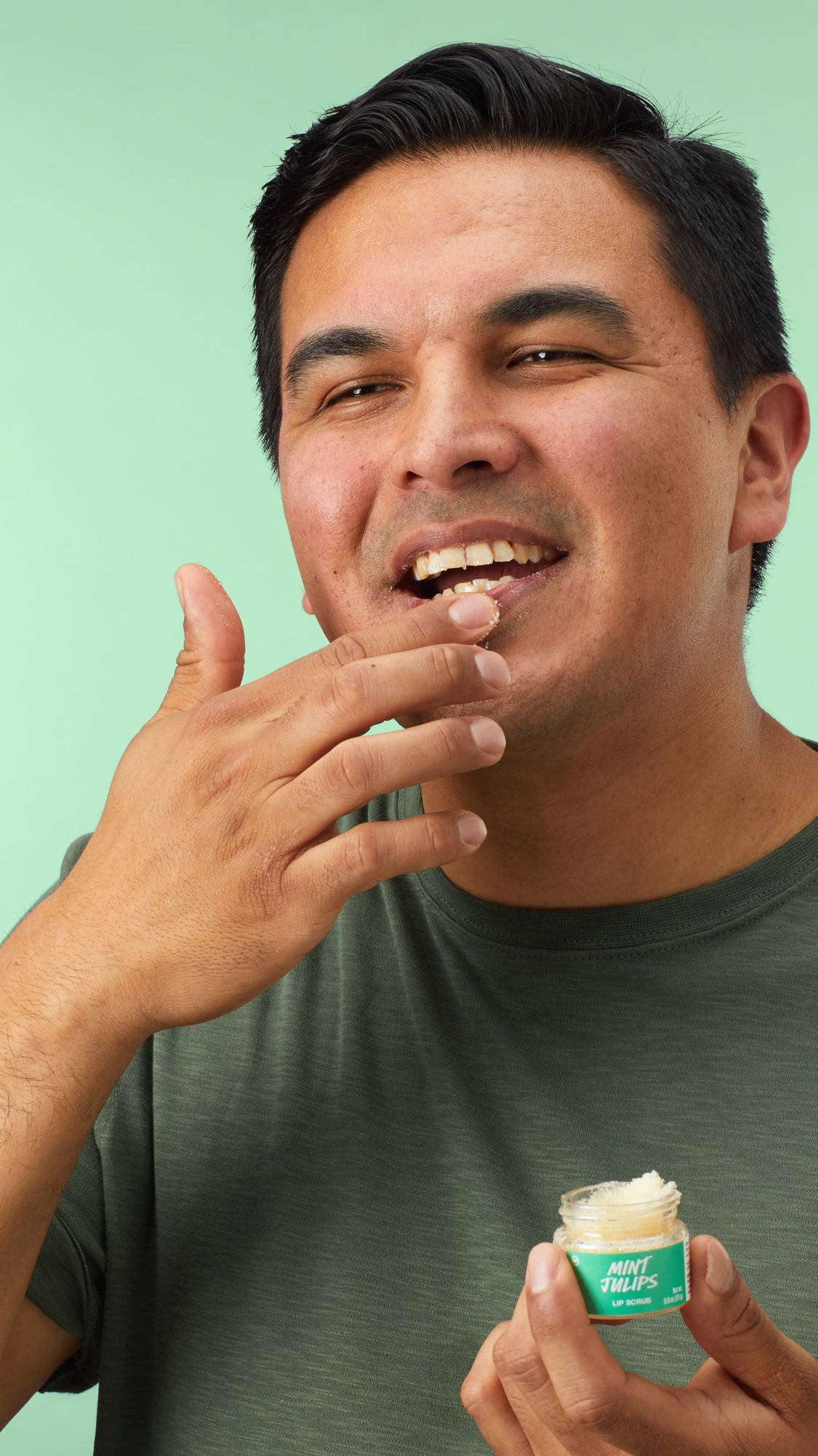 The image shows a model in a green t-shirt smiling as they apply the Lush 'Mint Julips' lip scrub to their lips with their finger. They hold the small glass jar in their other hand against a plain mint green background. 