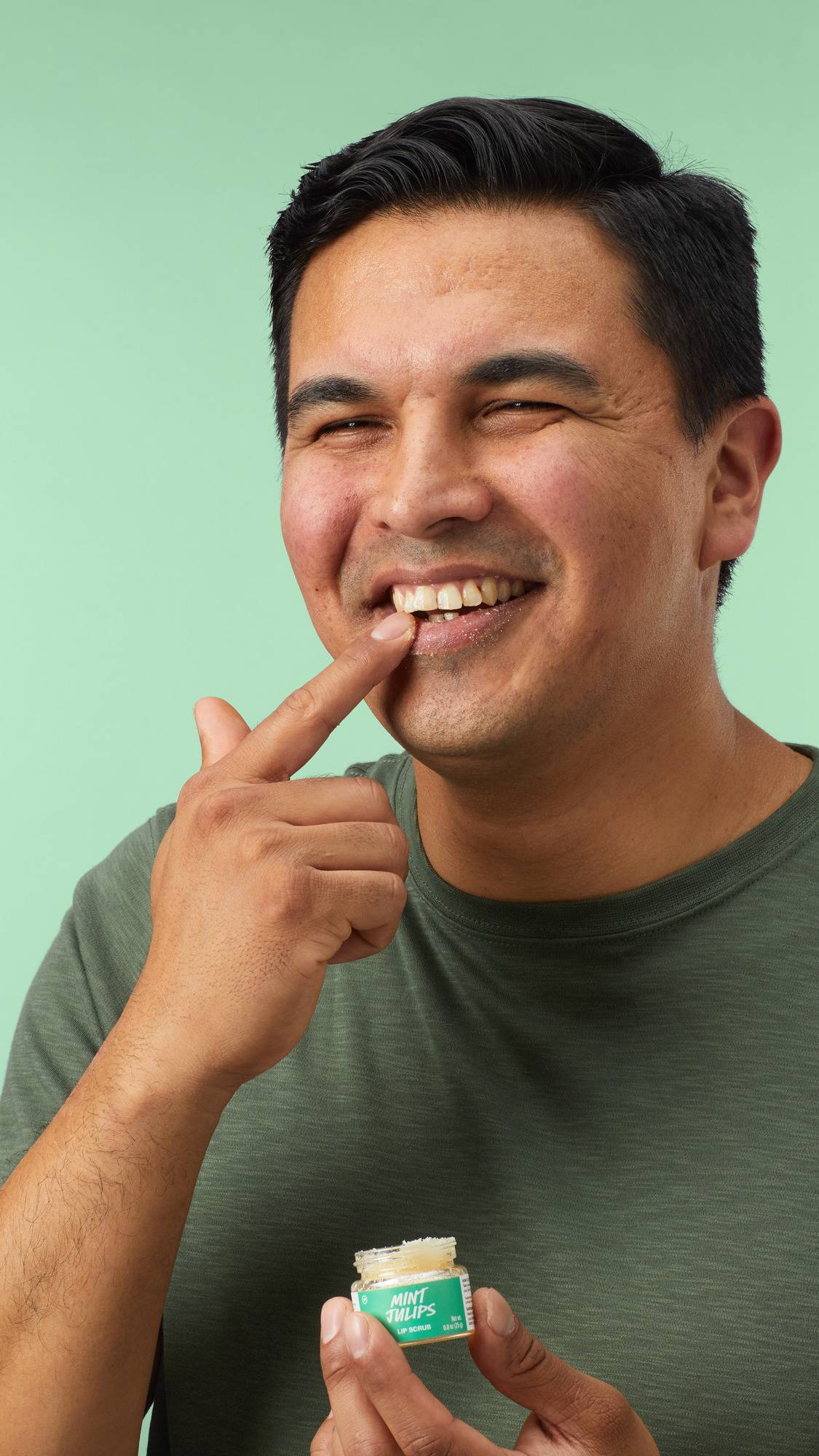 The image shows a model in a green t-shirt smiling as they apply the Lush 'Mint Julips' lip scrub to their lips with their finger. They hold the small glass jar in their other hand against a plain mint green background. 