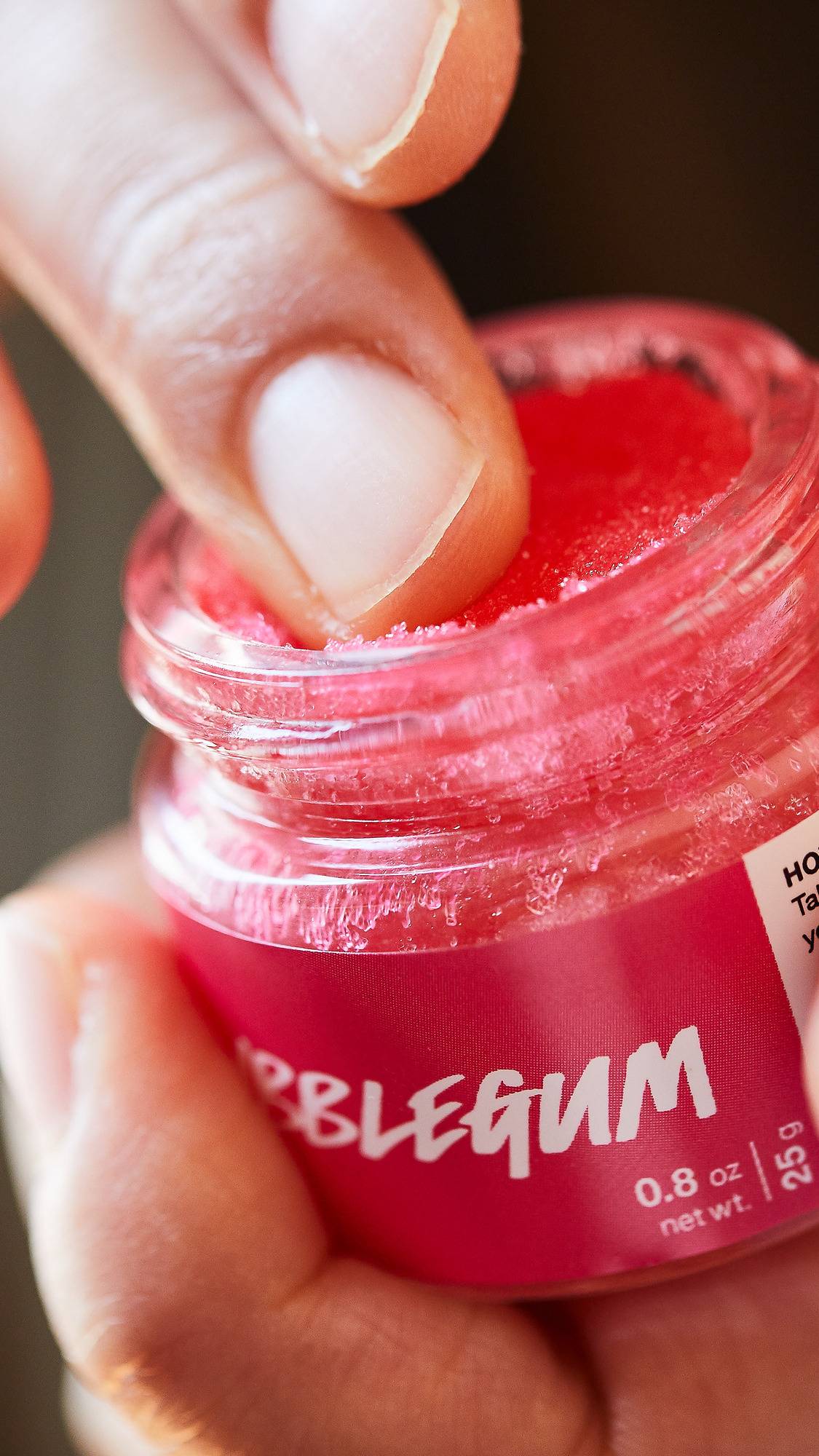  A close-up shot of a person's finger dipping into a small glass jar of Lush 'Bubblegum' lip scrub. The scrub has a vibrant pink, grainy sugar texture. The jar features a bright pink label with white text. 