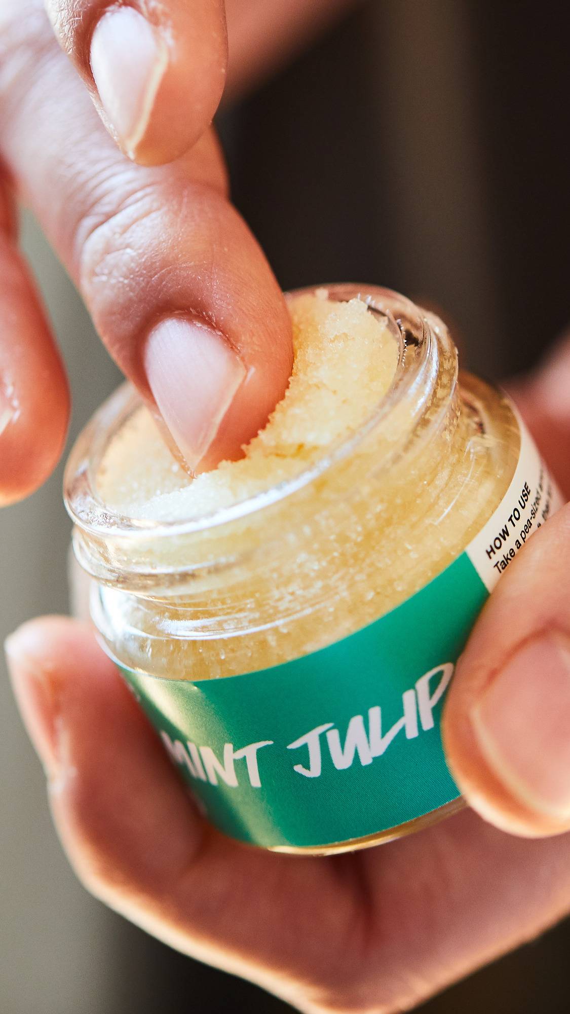 A close-up shot of a person's finger dipping into a small glass jar of Lush 'Mint Julips' lip scrub. The scrub has a pale yellow, grainy sugar texture. The jar features a bright green label with white text. 