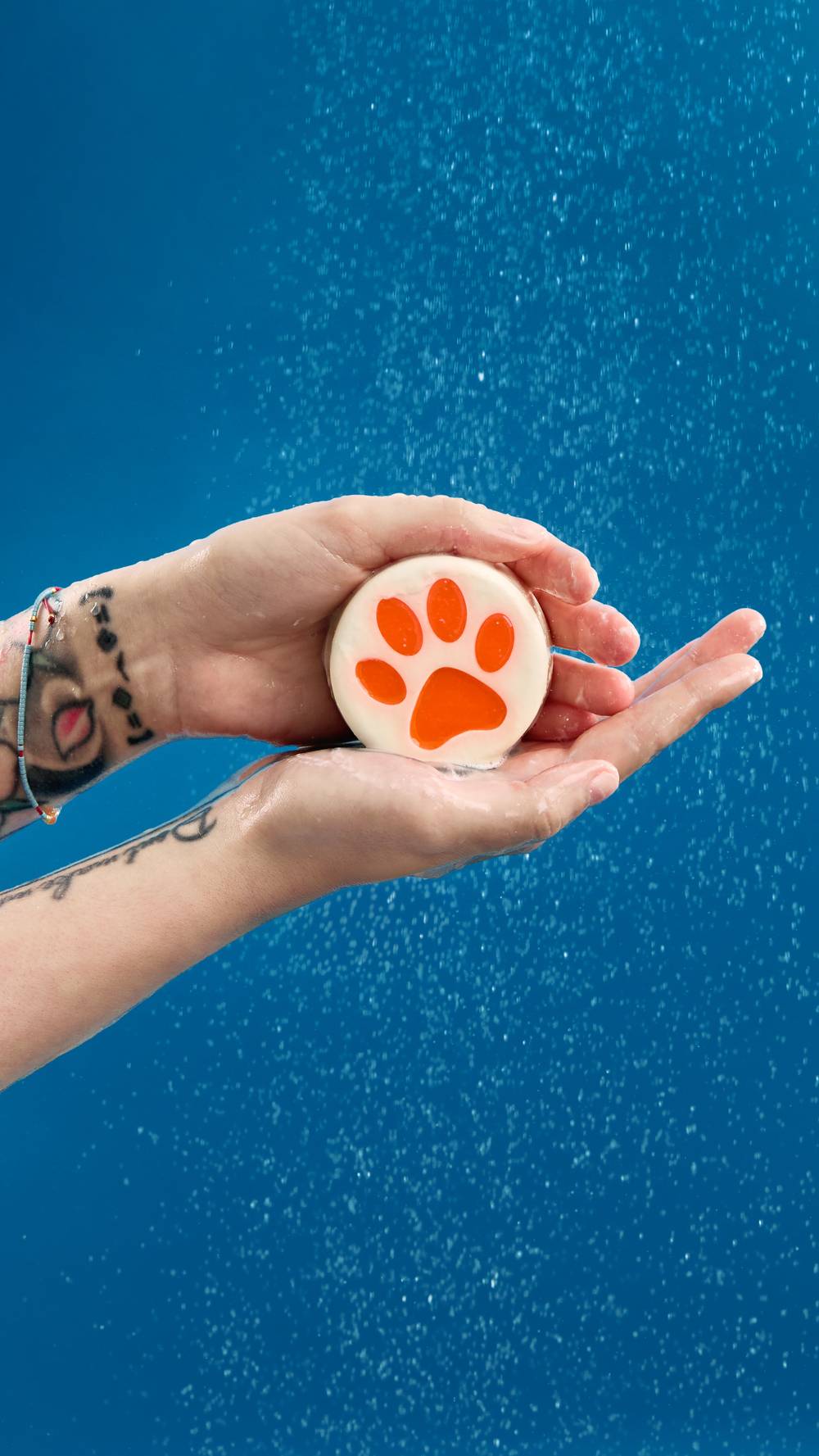Two cupped, wet hands with wrist tattoos hold a round white soap bar featuring a bright animal paw print design. Water droplets spray all around the hands against a plain, bold blue background.