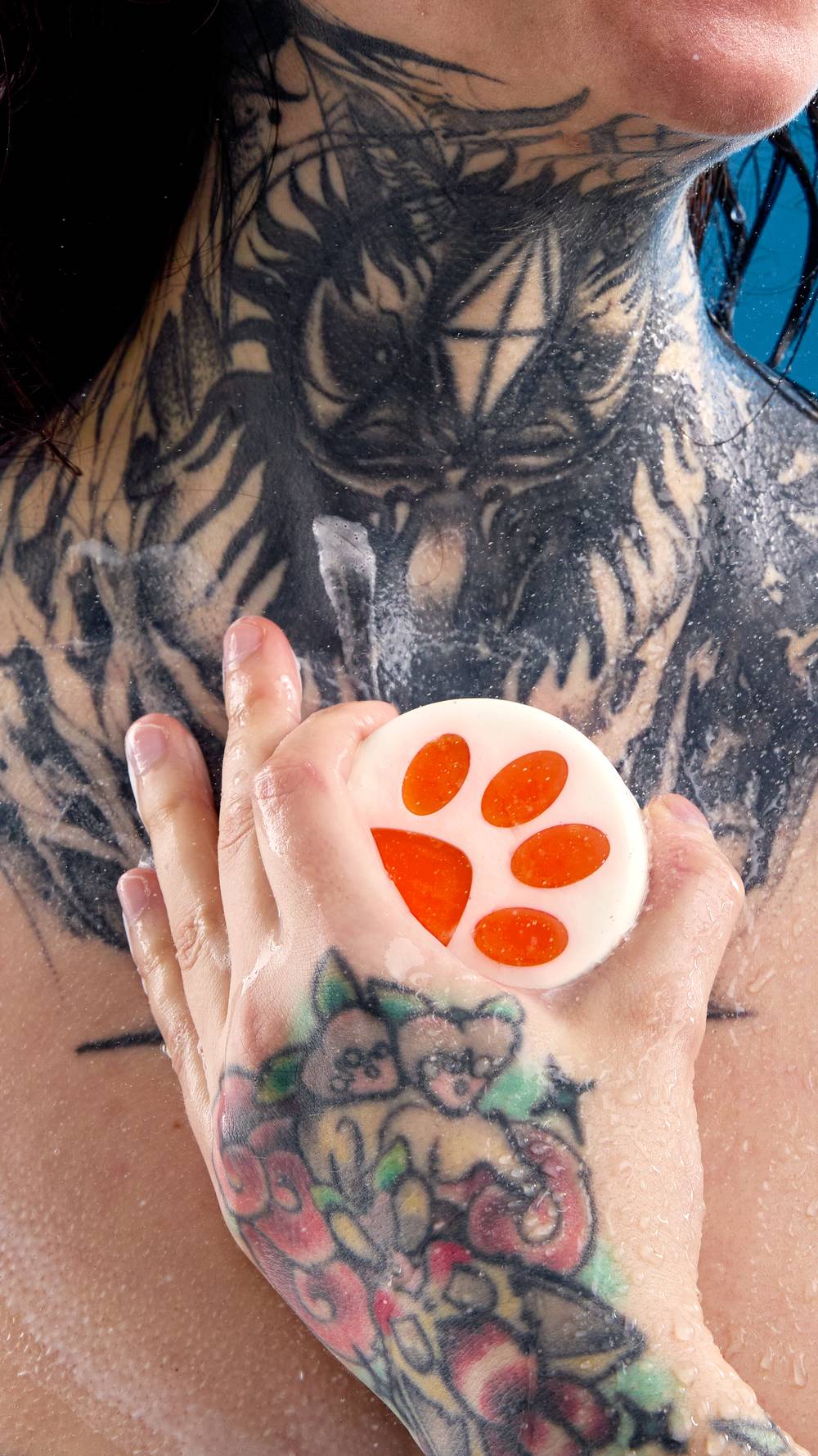 A close-up shows a tattooed hand holding a round white bar of soap with an orange paw print design. The soap is being lathered against a wet, heavily tattooed chest and neck in the shower.