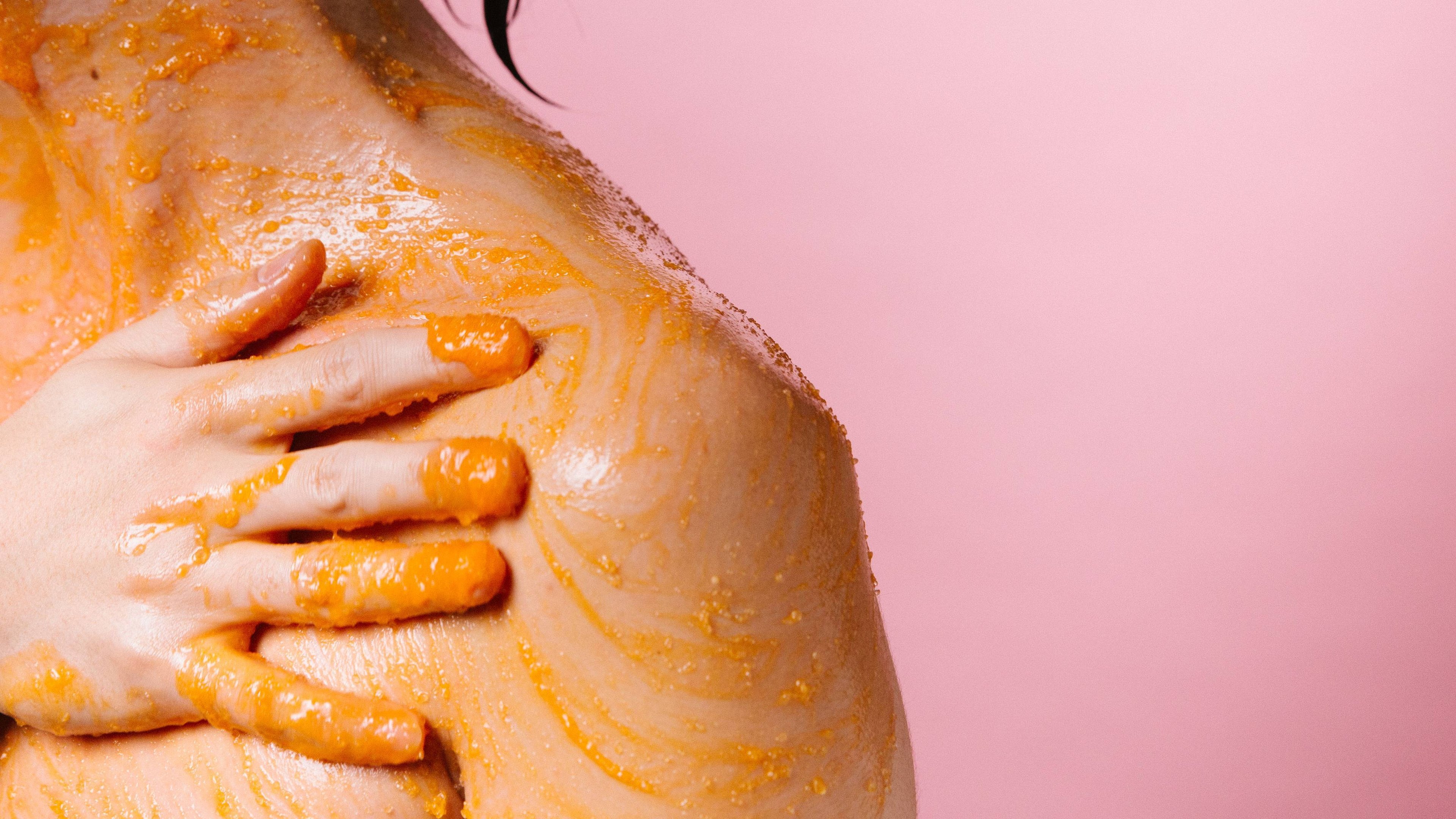 A close-up image of the model's bare shoulder on a pastel-pink background. They are applying the Orange body scrub to their skin and exfoliating in circular motions.
