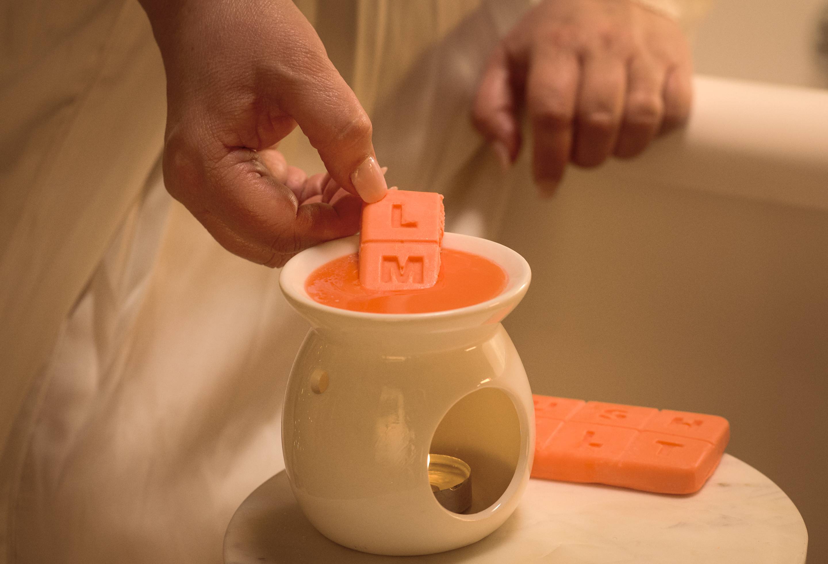 The image shows a close-up of the model placing two pieces of wax melt into a white wax burner with a lit tealight. 